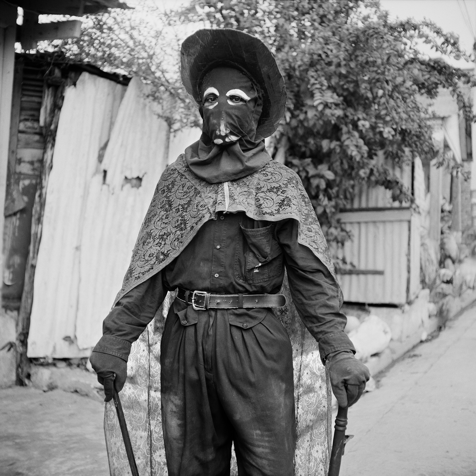 A man wearing a lace cape, cloth mask and cardboard hat with gloves.