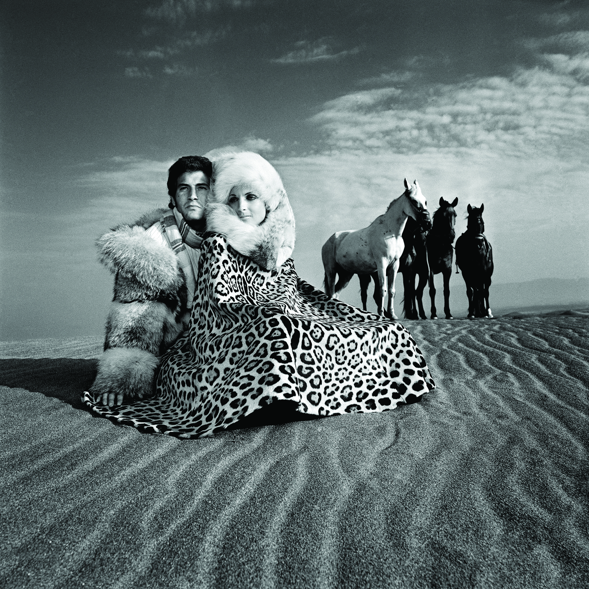 black-and-white image of a fur-wearing couple in the desert in front of four horses