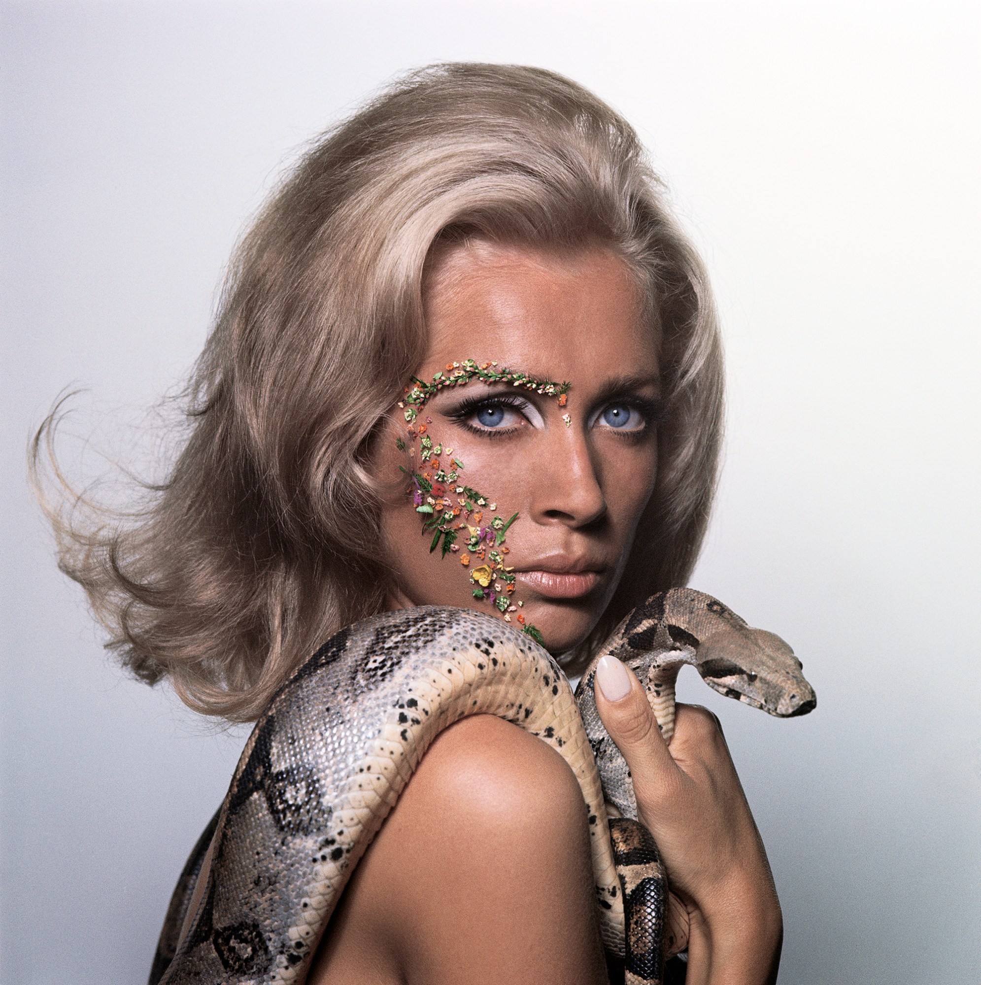 a blonde woman seen in profile holding a snake over her shoulder wearing gems on one side of her face