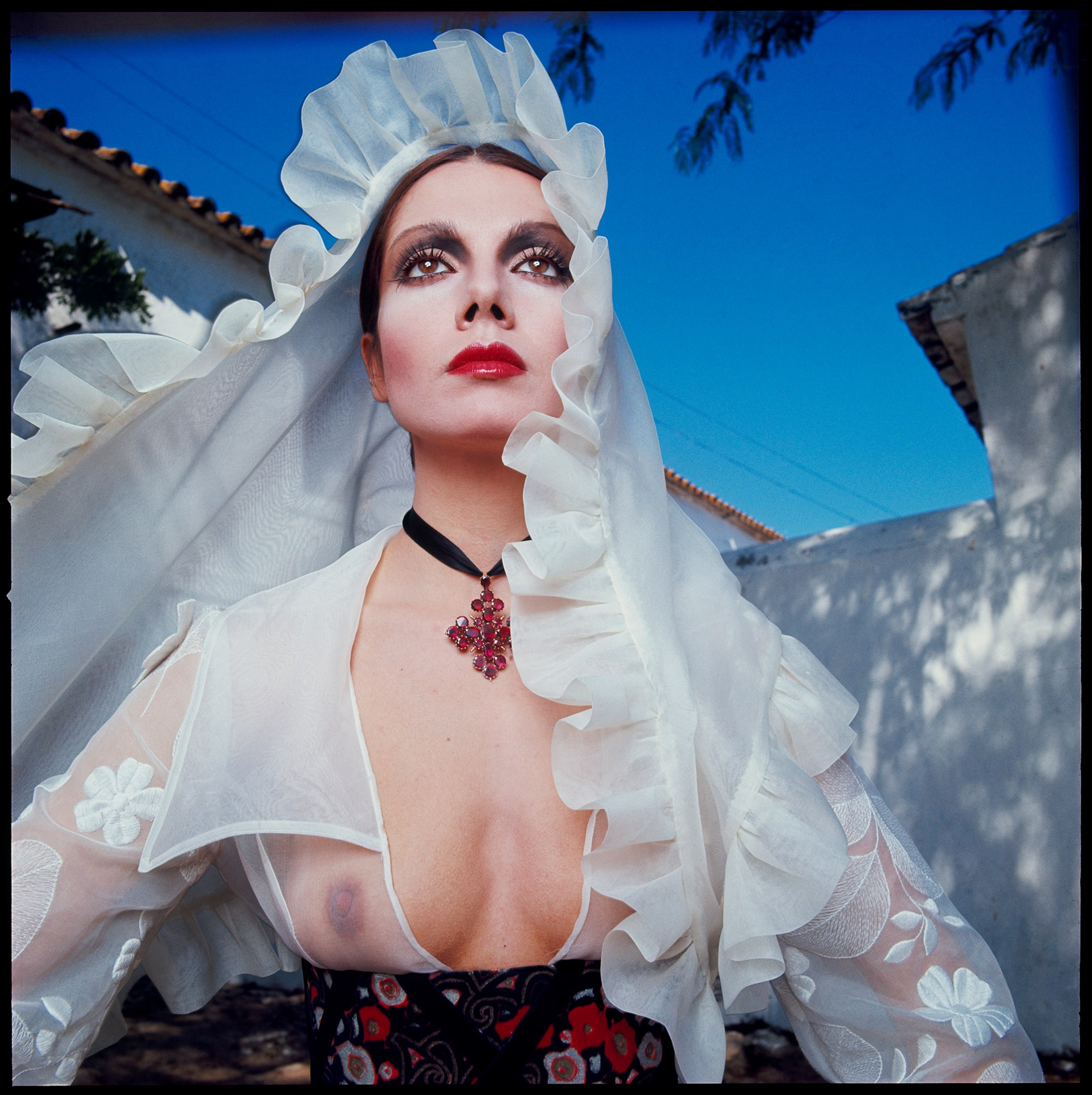 a woman in translucent white shirt and veil, seen against a blue sky and white wall