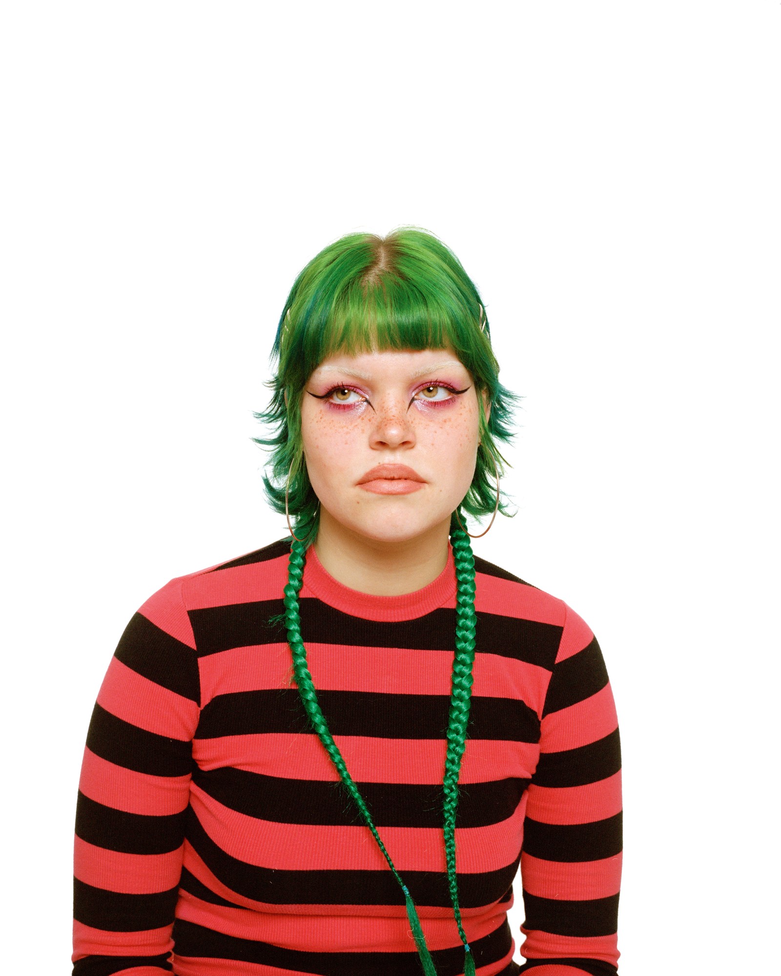 a person with bright green hair braids and a striped red and black top looks sullenly off-camera