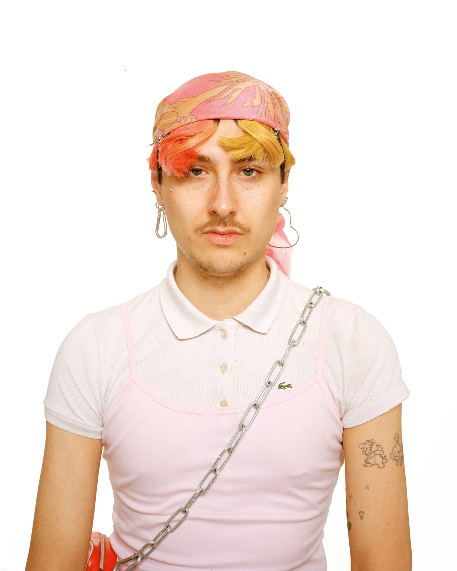 a person with colourful hair and a moustache poses in a pale pink polo look with chain bag