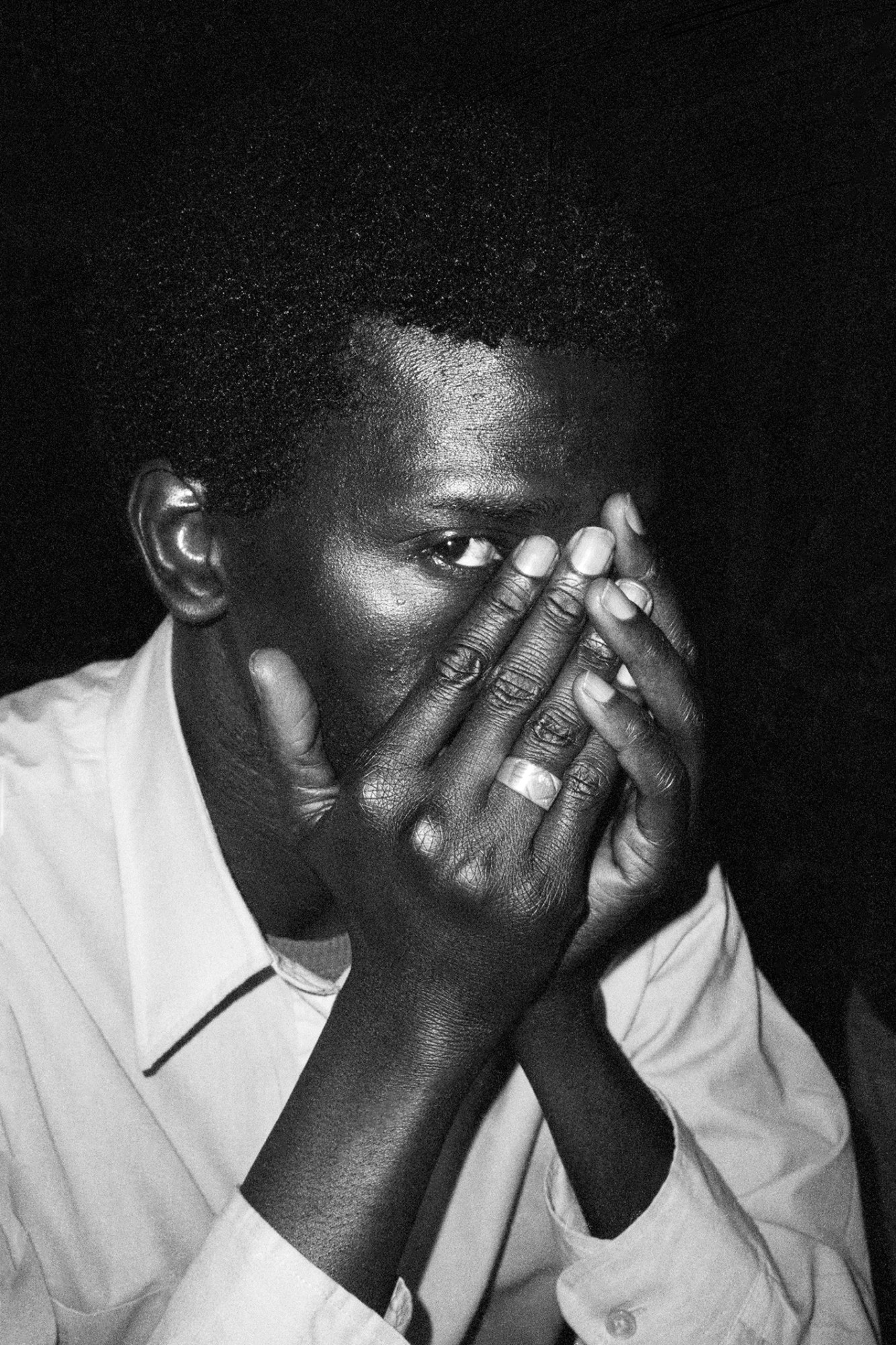 a dark-skinned Black man in a light shirt peers out from behind his hands, he wears a ring on one finger