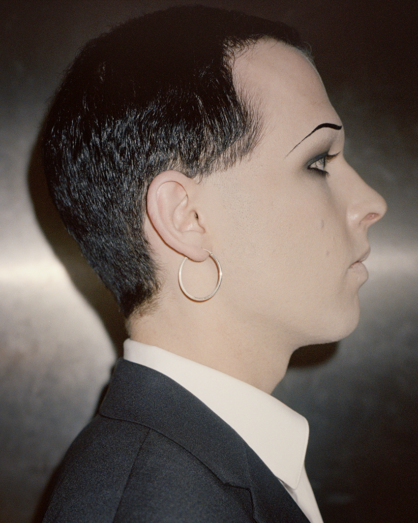 a side profile of person in a suit and hoop earrings; they wear smoky eye makeup and a thin drawn-on eyebrow