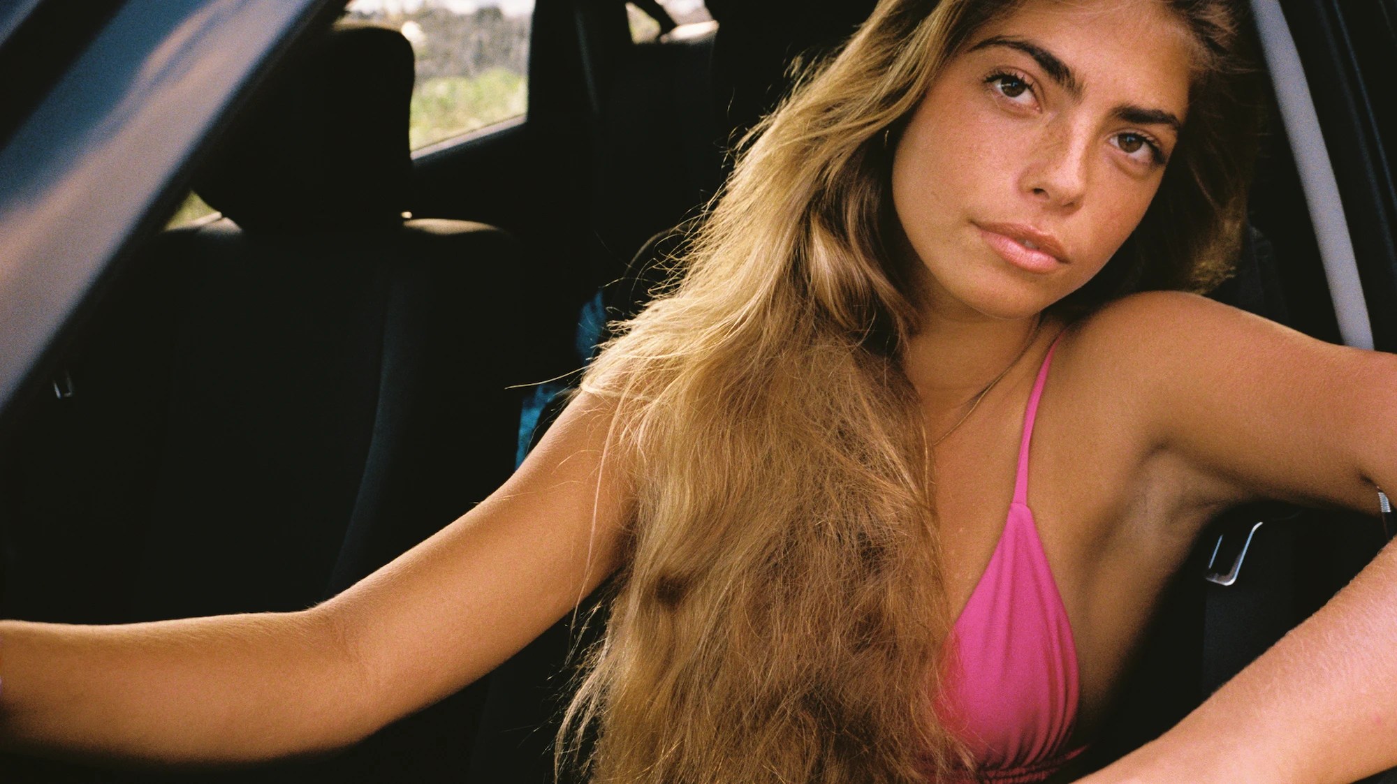 a woman with long brown hair wearing a pink bikini sits in the front seat of a car, looking at the camera out of the open window