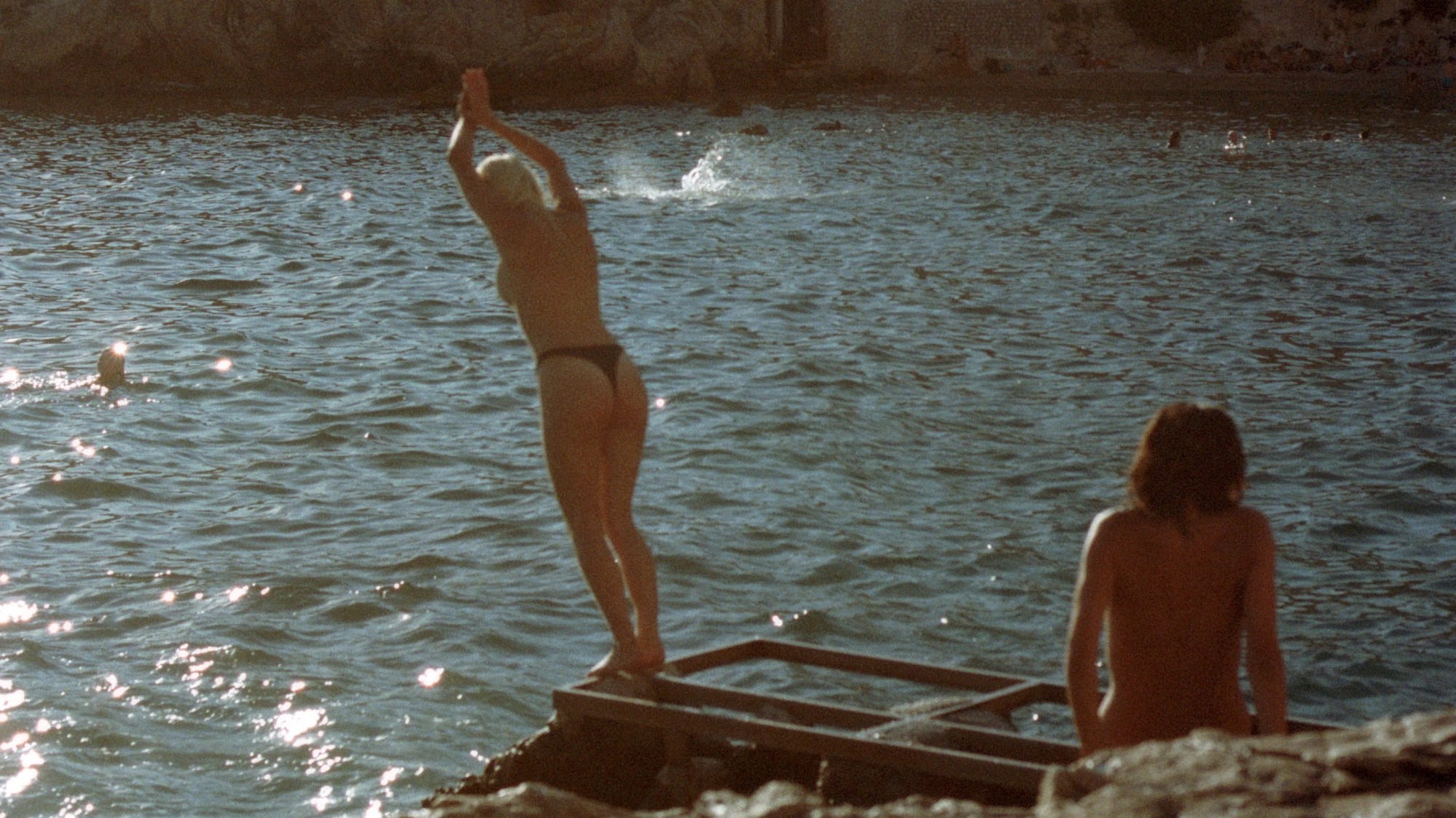 two naked people on a metal pier. one has their hands outstretched ready to jump in the water. the other lounges on the shore.