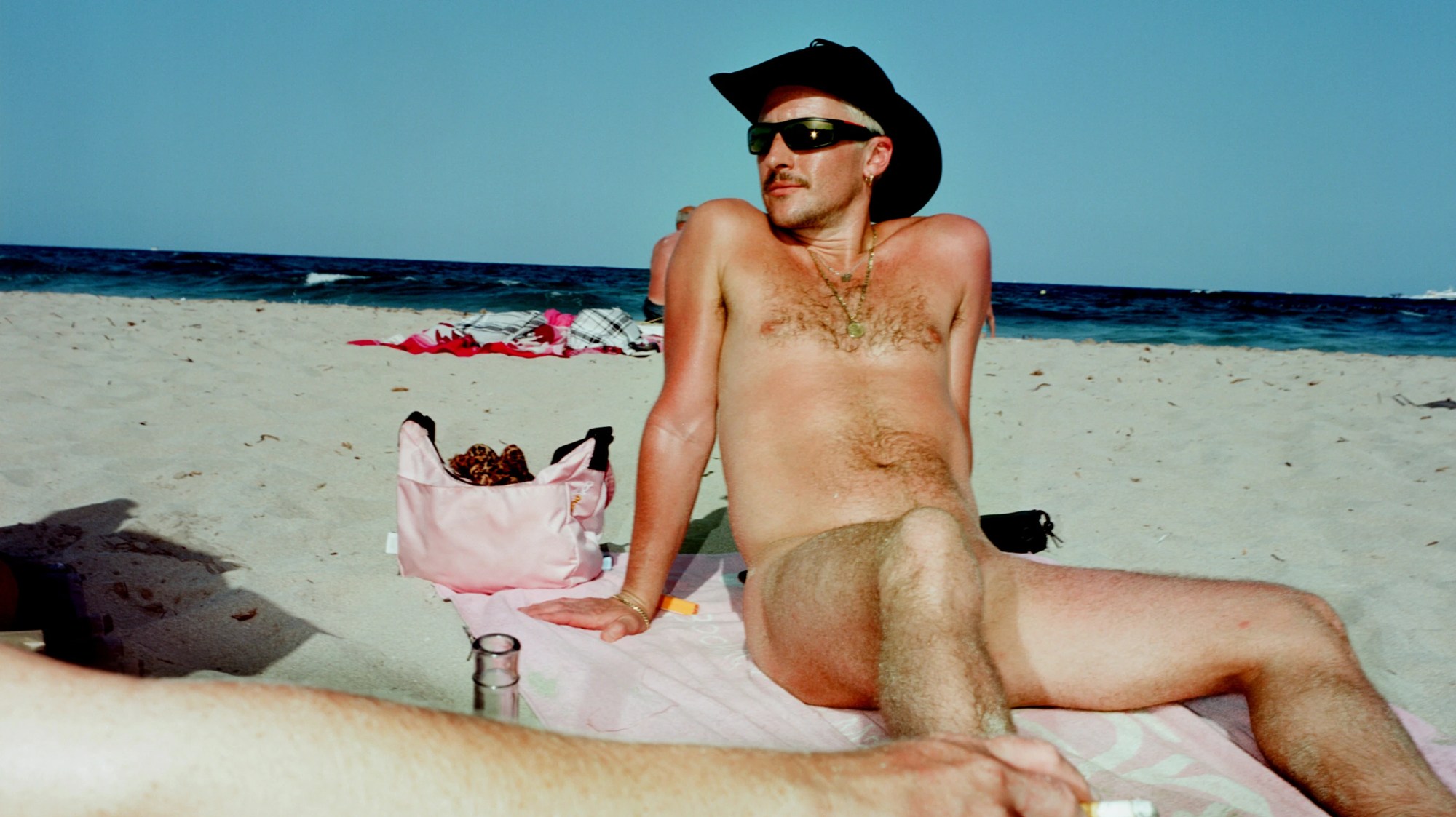 a man naked on a beach, his knee covering his crotch. the sky is blue. he's wearing a cowboy hat and wraparound sunglasses