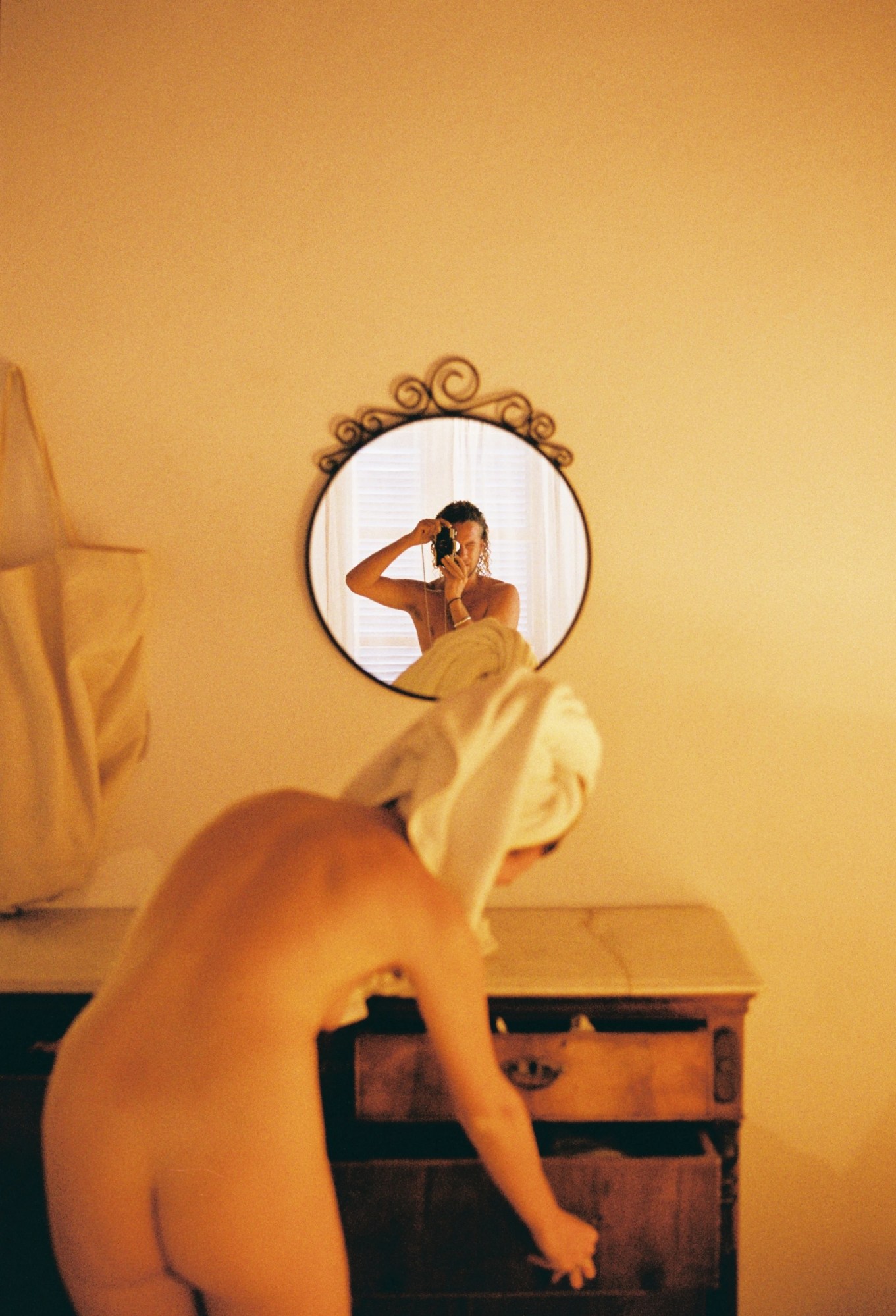 a man with wet hair takes a photo in the mirror, a woman with her hair in a towel but otherwise completely naked is in view, her naked body seen from behind.