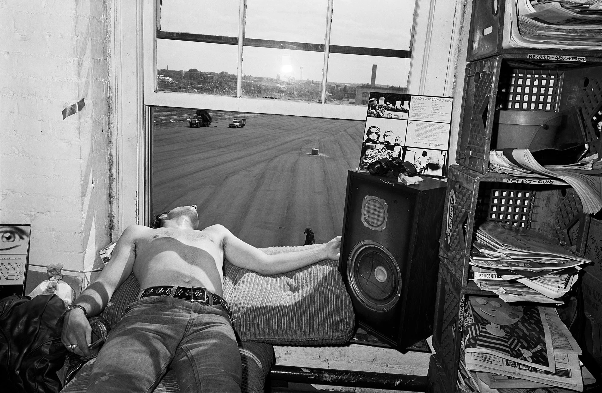 a man wearing jeans and no shirt leans bacwards out of a bedroom window, with a speaker by the window sill.