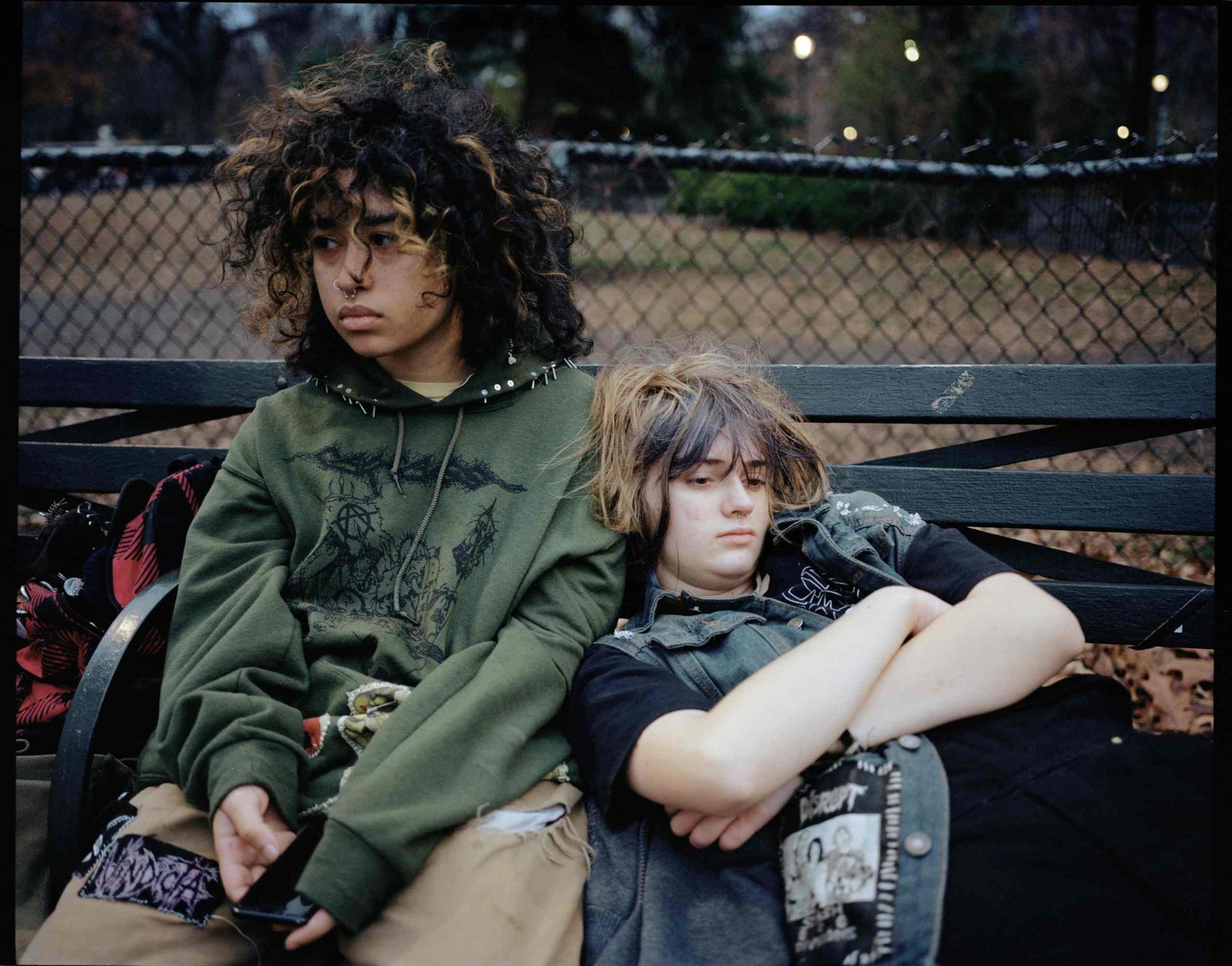 two bored looking teenagers dressed in skatewear with long hair sit on a bench. one rests on the others shoulder.