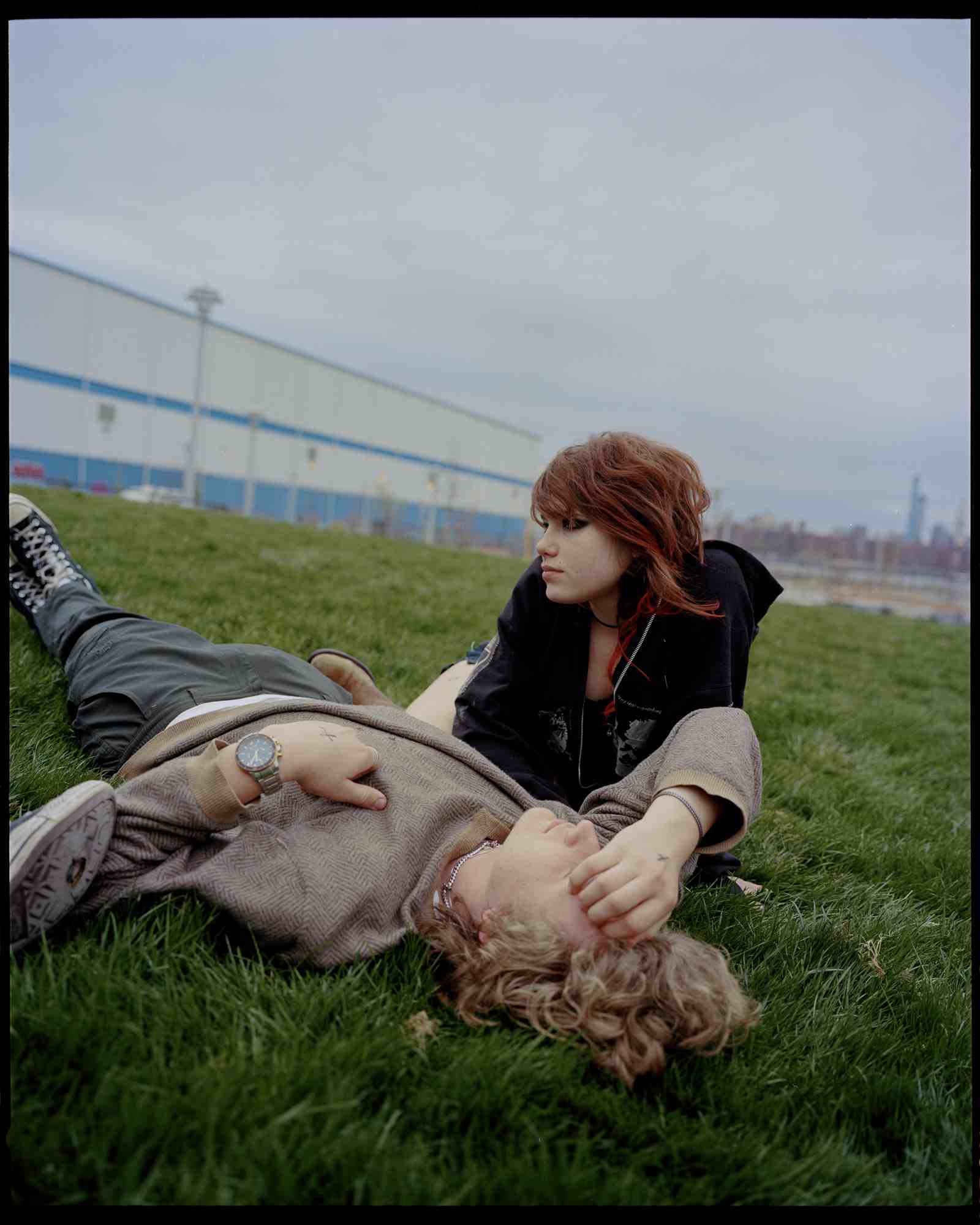 two teenagers lie on grass. one boy with blonde hair lies looking at the sky. another looks off camera, a girl wearing heavy eye make-up.