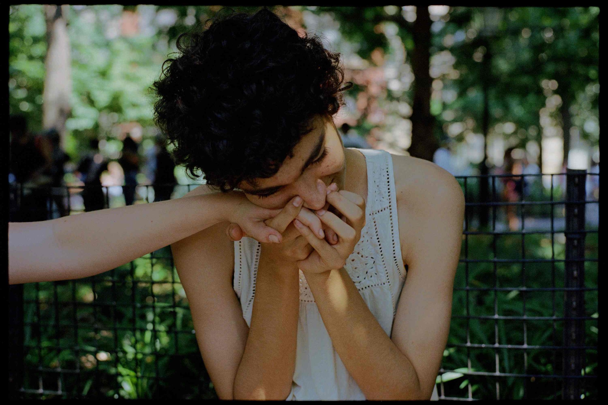 a teenager in a crocheted style white vest kisses the hand of a friend off camera. they have curly hair and look happy.