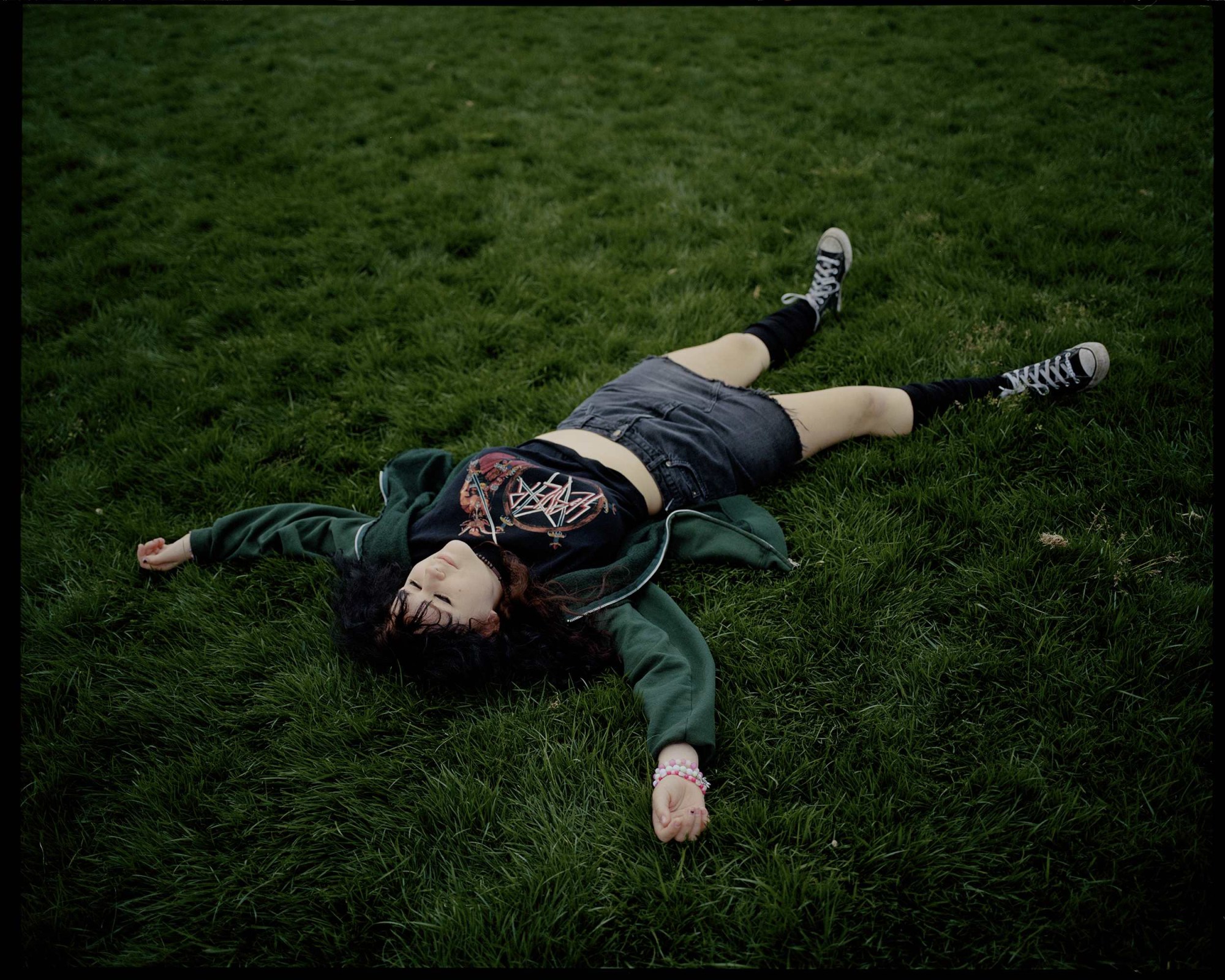 a teenager with a slayer crop top and denim skirt lies on the grass and looks up at the sky.