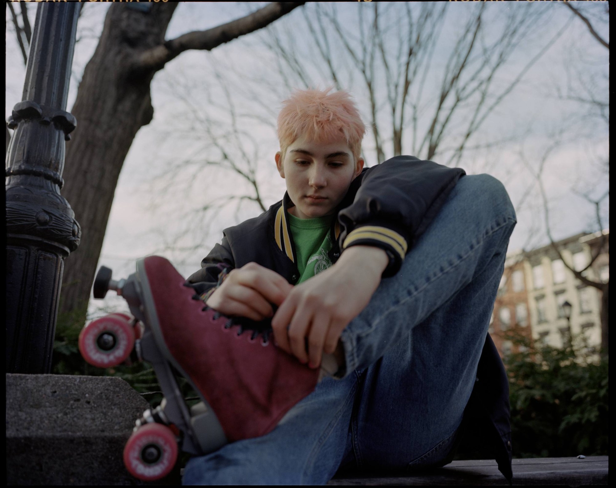 a teenager with short pastel pink hair laces up a red roller skate on a bench.