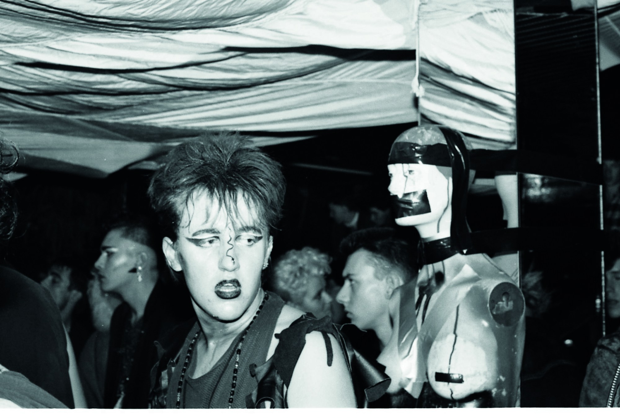 a person with goth meets new romantic makeup and hair standing by a taped-up mannequin in a club