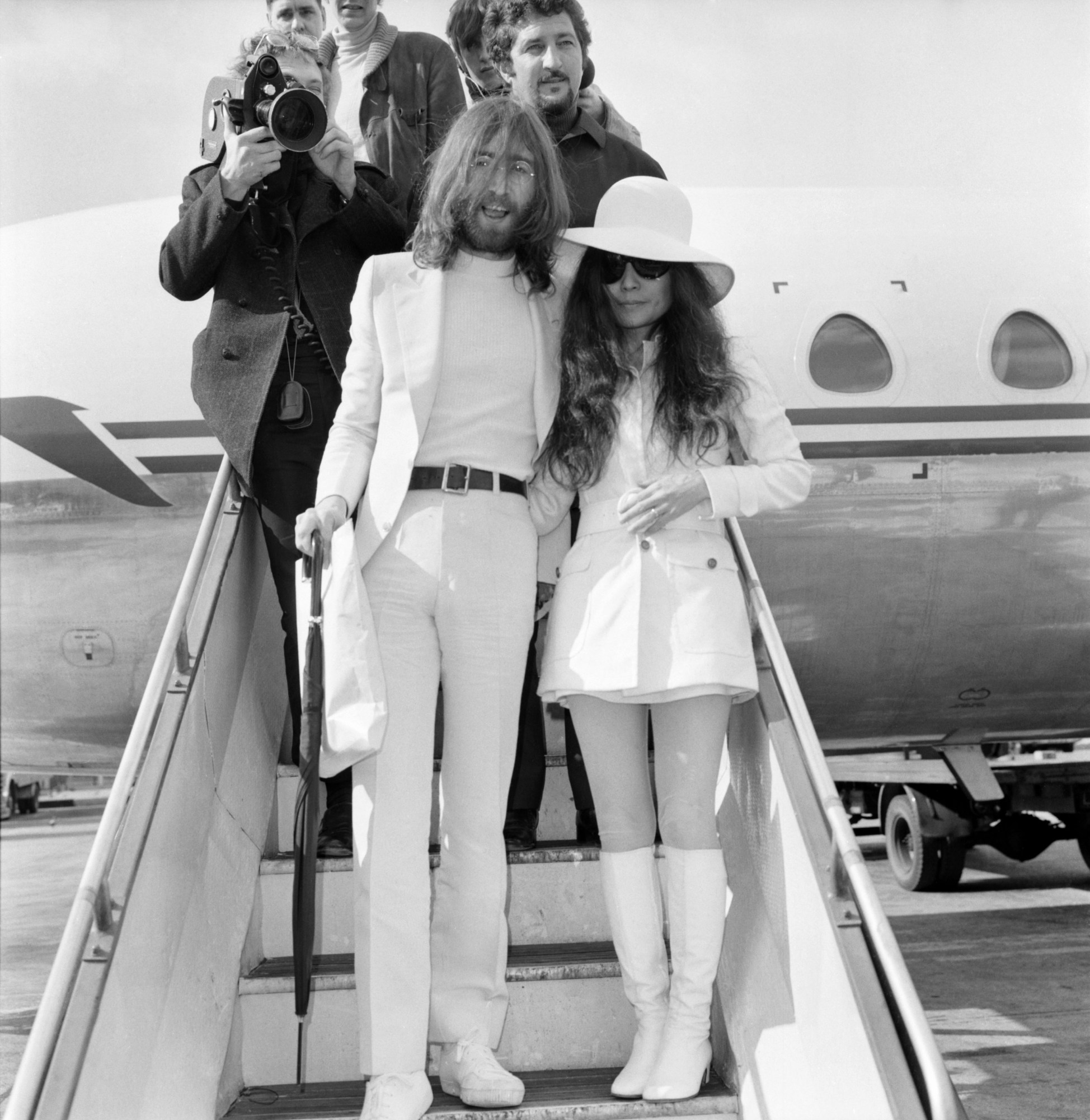 john lennon and yoko ono standing on the steps of a plane wearing all white. their team, including a man with a film camera, are behind them exiting the plane