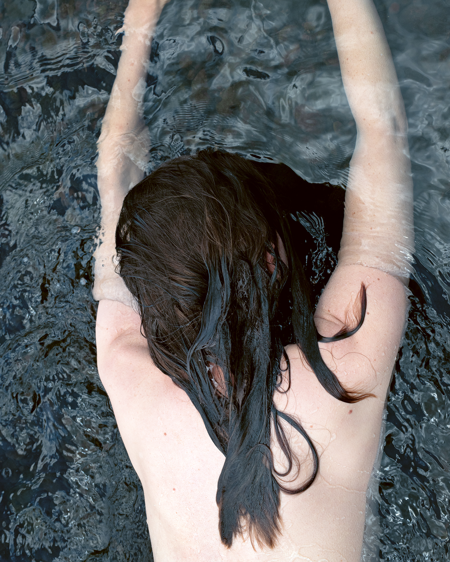 a woman lies naked face down in water that covers her arms and wets her hair