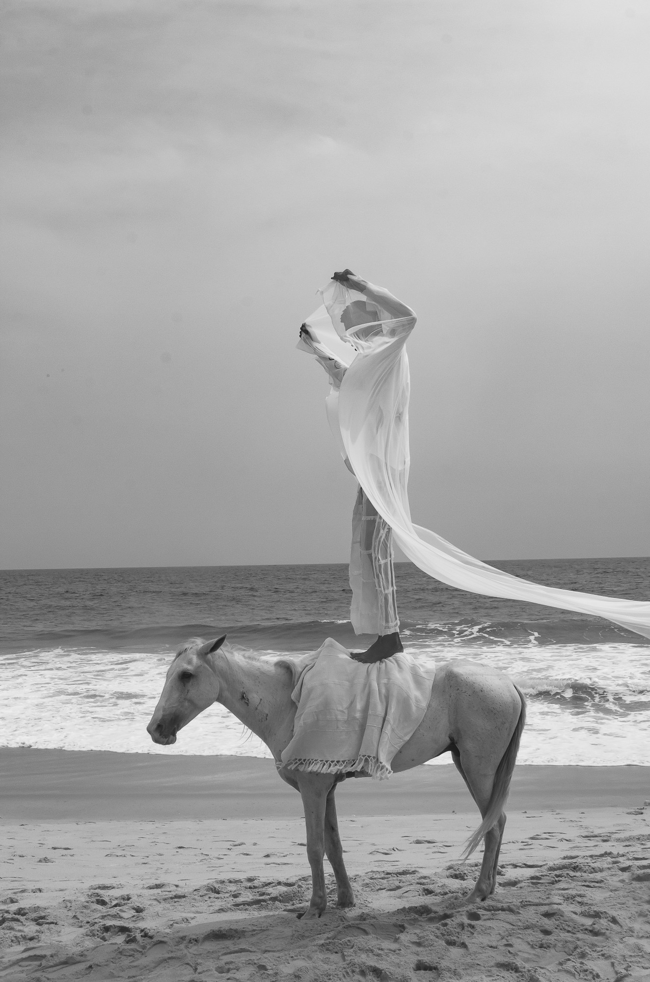 A man draped in white fabric standing on a horse on the beach.