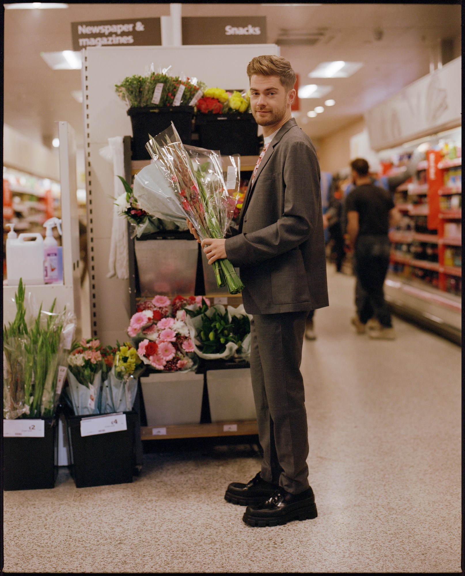 a portrait of male film director lukas dhont standing in a supermarket holding flowers. he has quiffed brown hair. he wears a grey suit and a patterned t shirt and looks into the camera