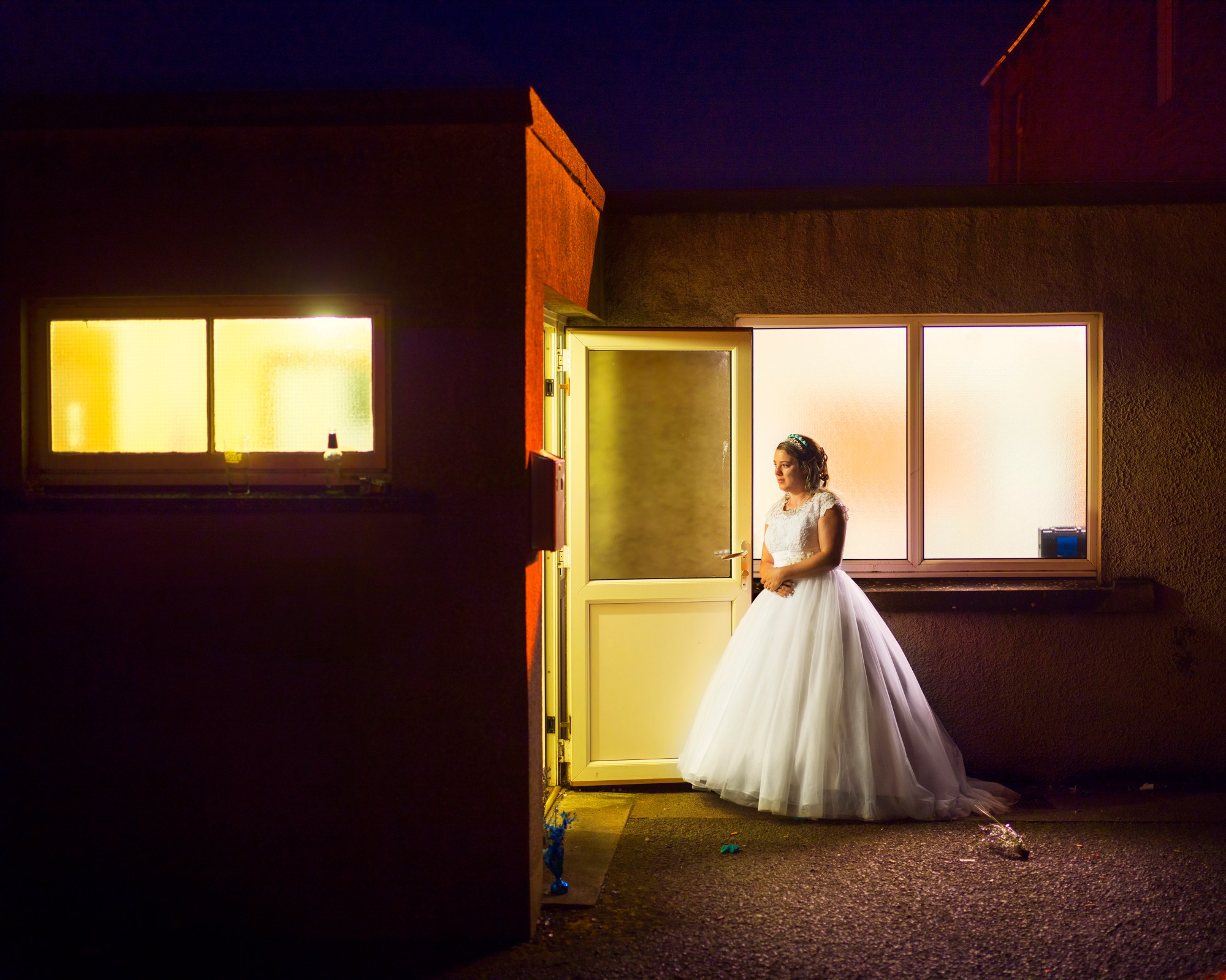 a woman in a large wedding dress stands outside, bathed in warm light from an open PVC door