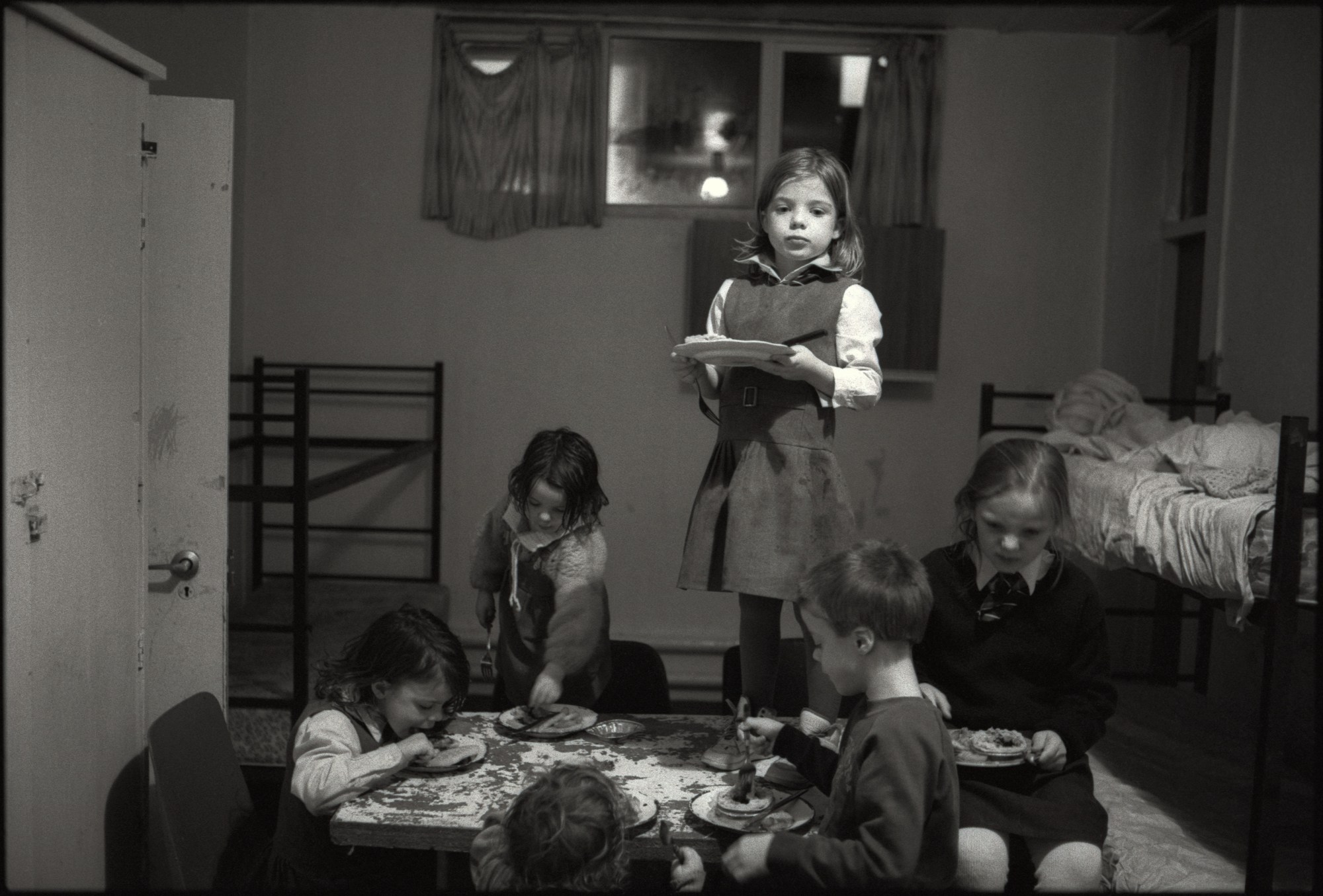 six young children in school uniforms eat their dinner in a room that hosts a dining table and a bed