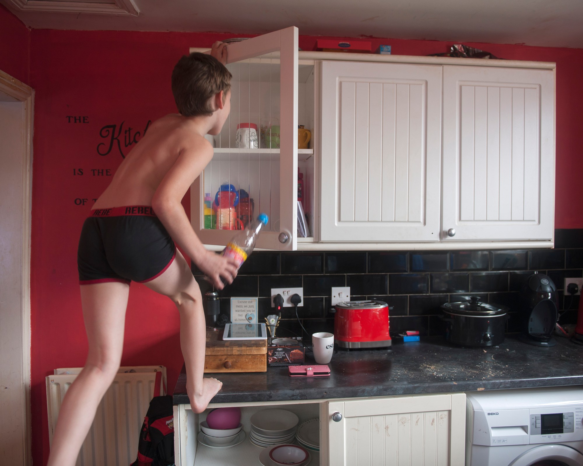 a young boy in boxers stands on the kitchen worktop taking a bottle of squash from a cupboard