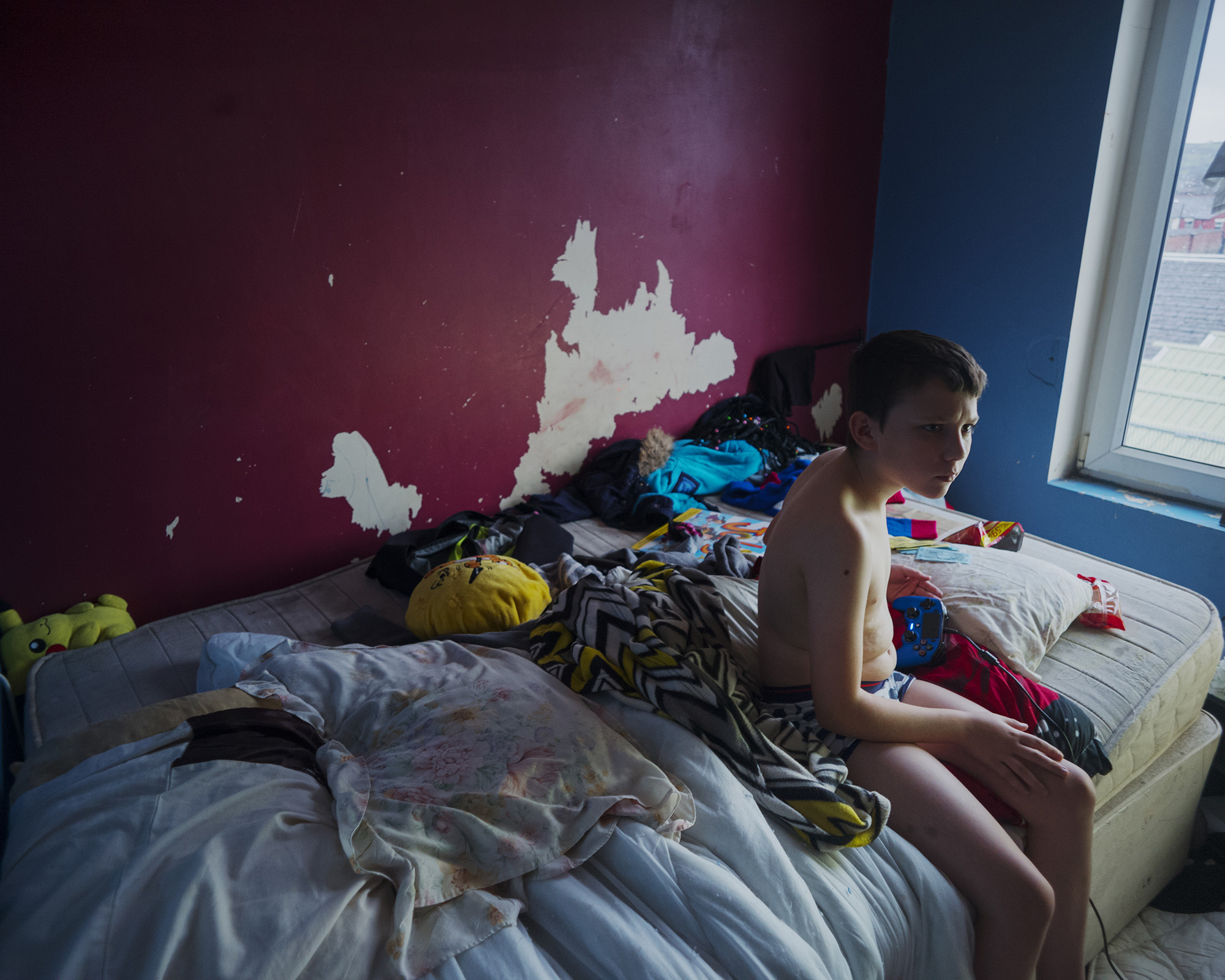 a boy sits in his pants on a messy bed; the red paint is flaking off the wall behind him