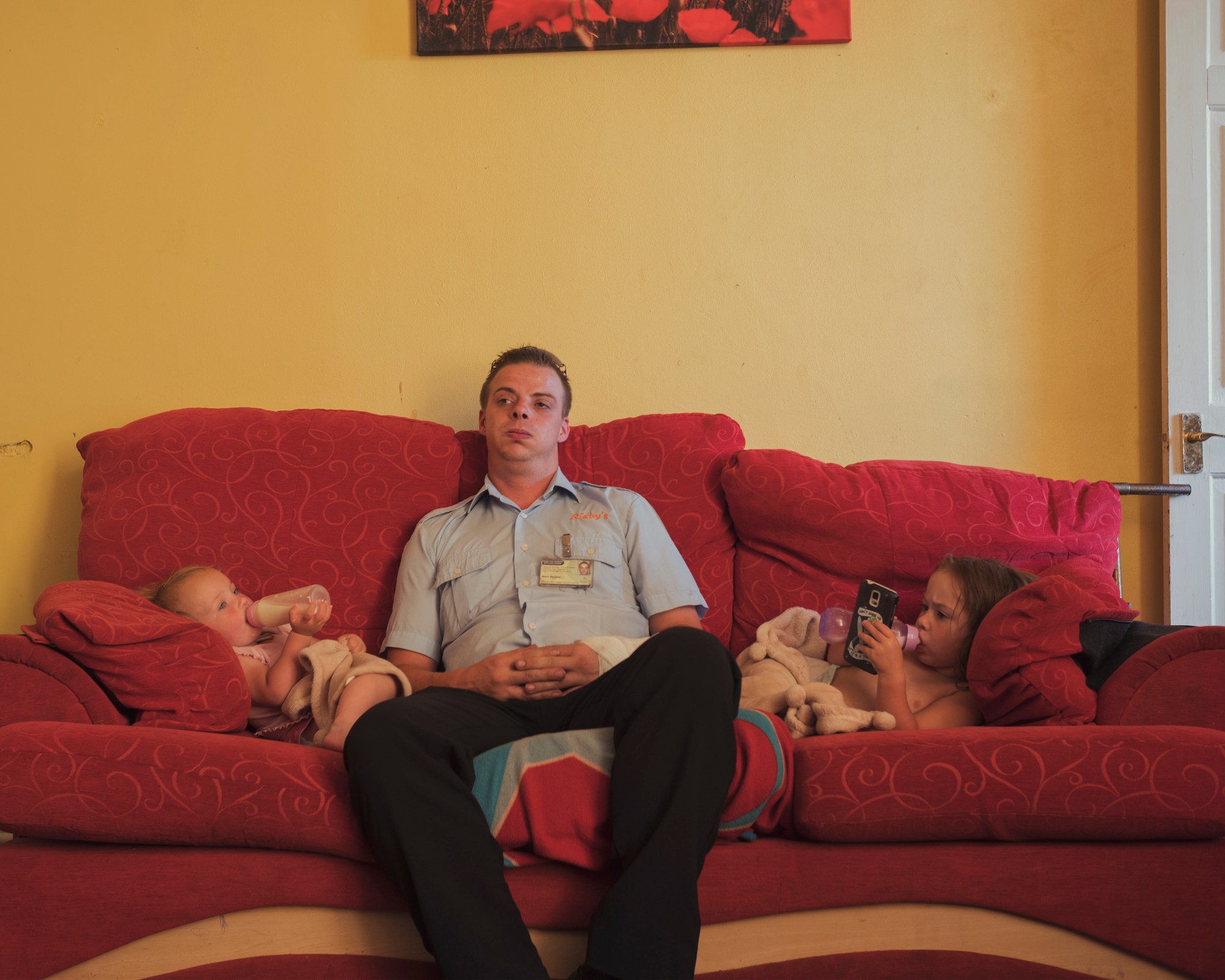 a man in uniform sits on the sofa between two young children drinking bottles of milk