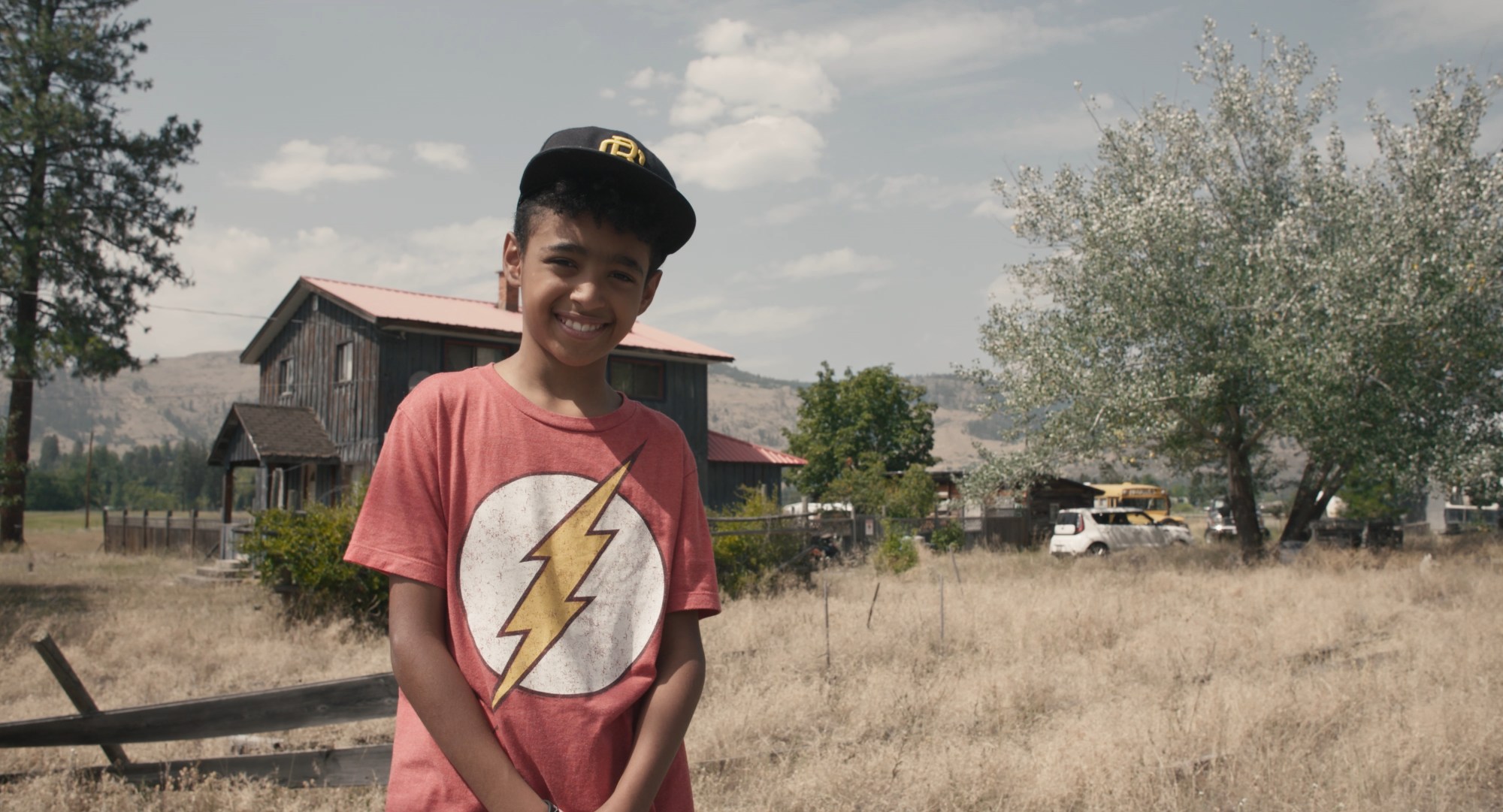 film still of a boy smiling in a red tshirt in front of his house