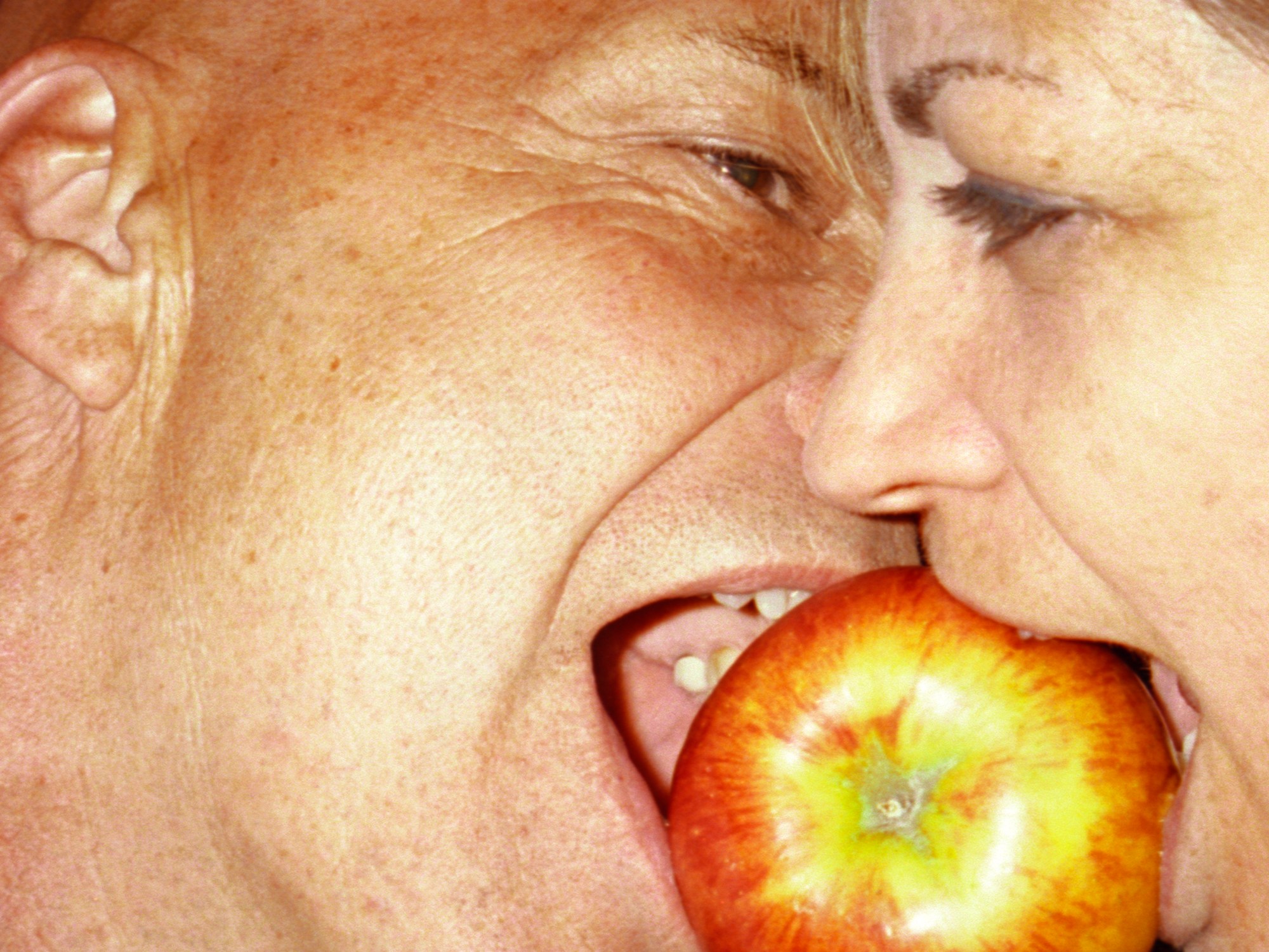 an older white couple both take a bite of the same apple in a close up shot