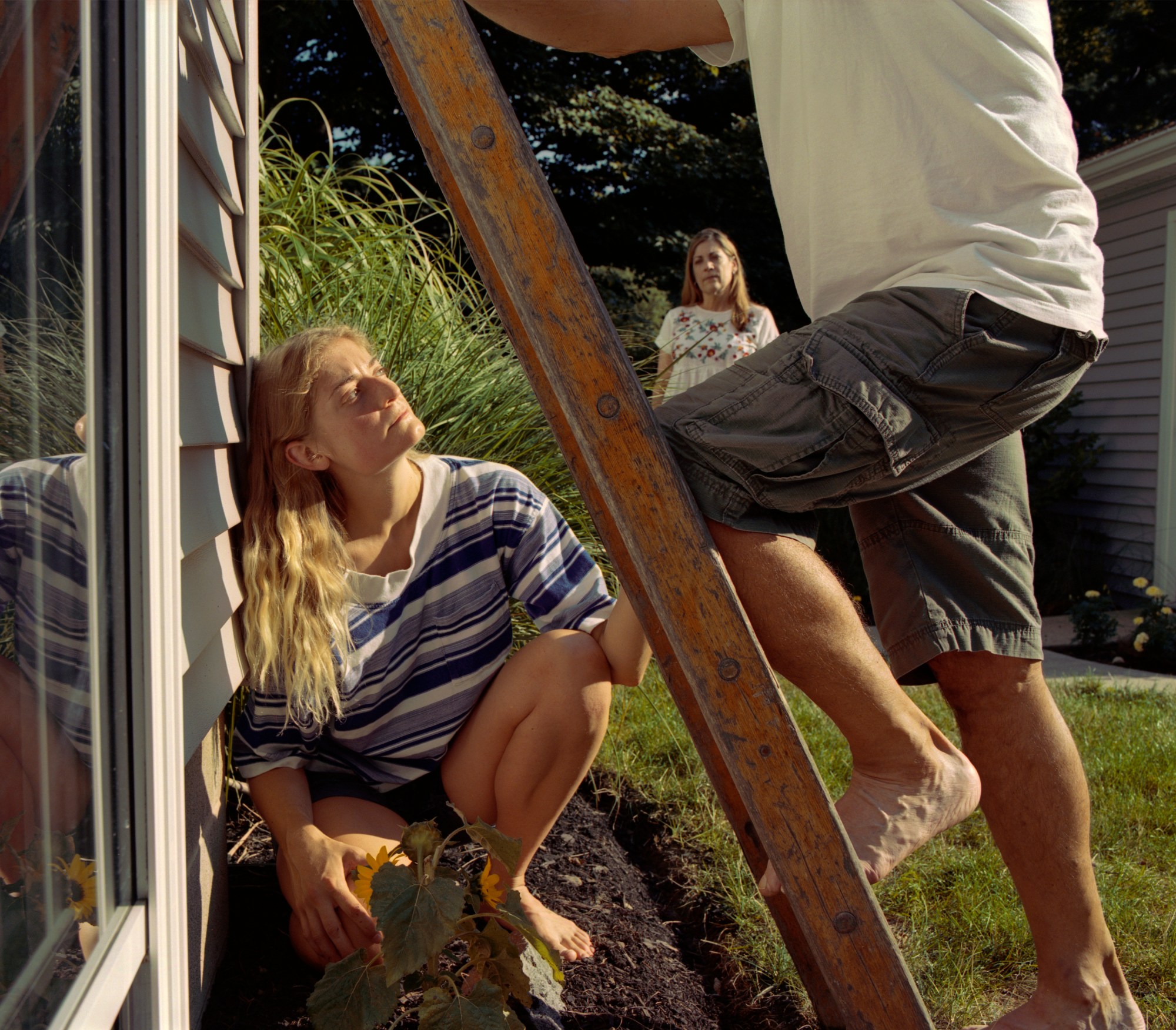 a young blond woman holds a latter as an older man climbs it. her mother watches on