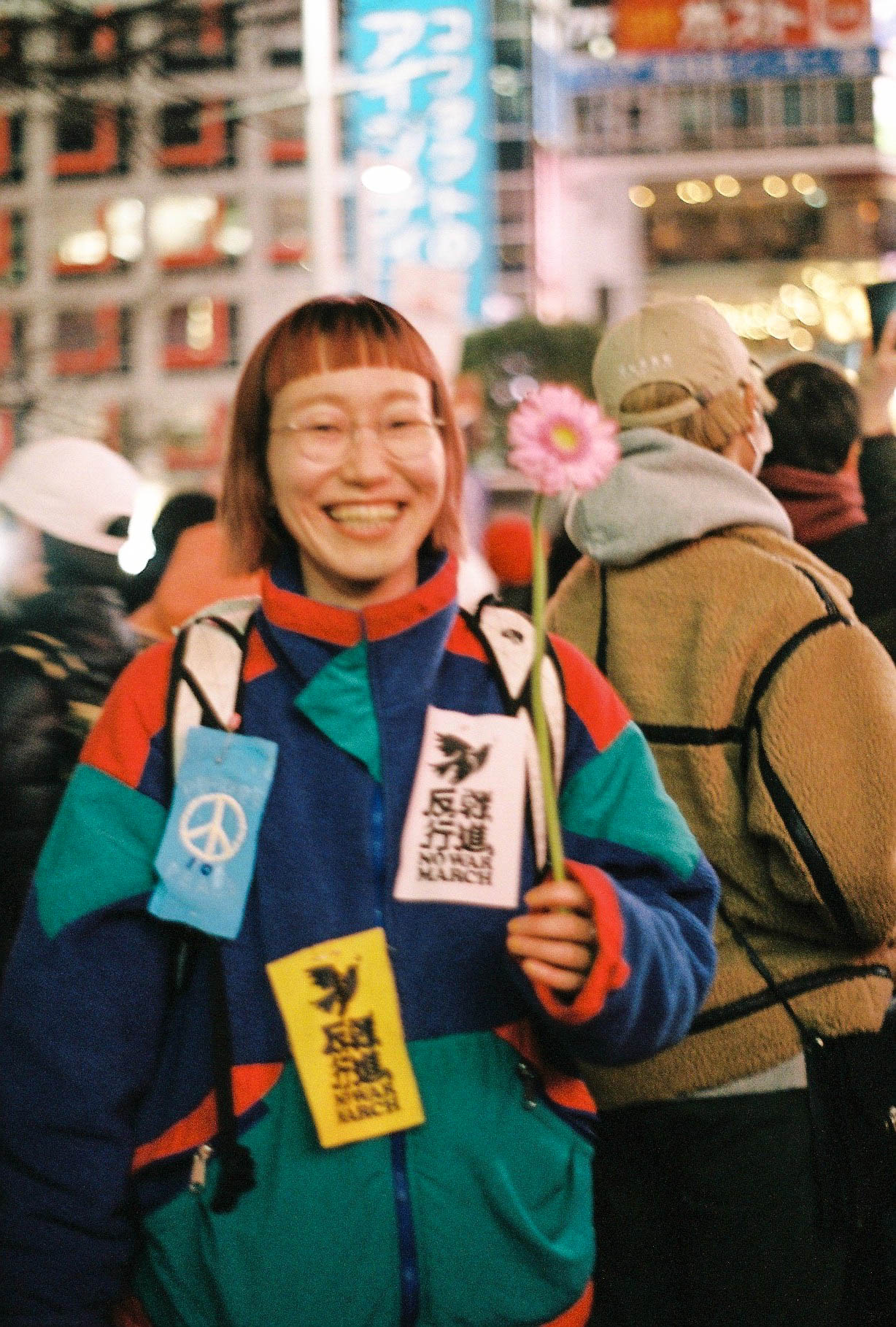 Photograph of a woman with a colourful coat and holding a flower standing amongst a crowd