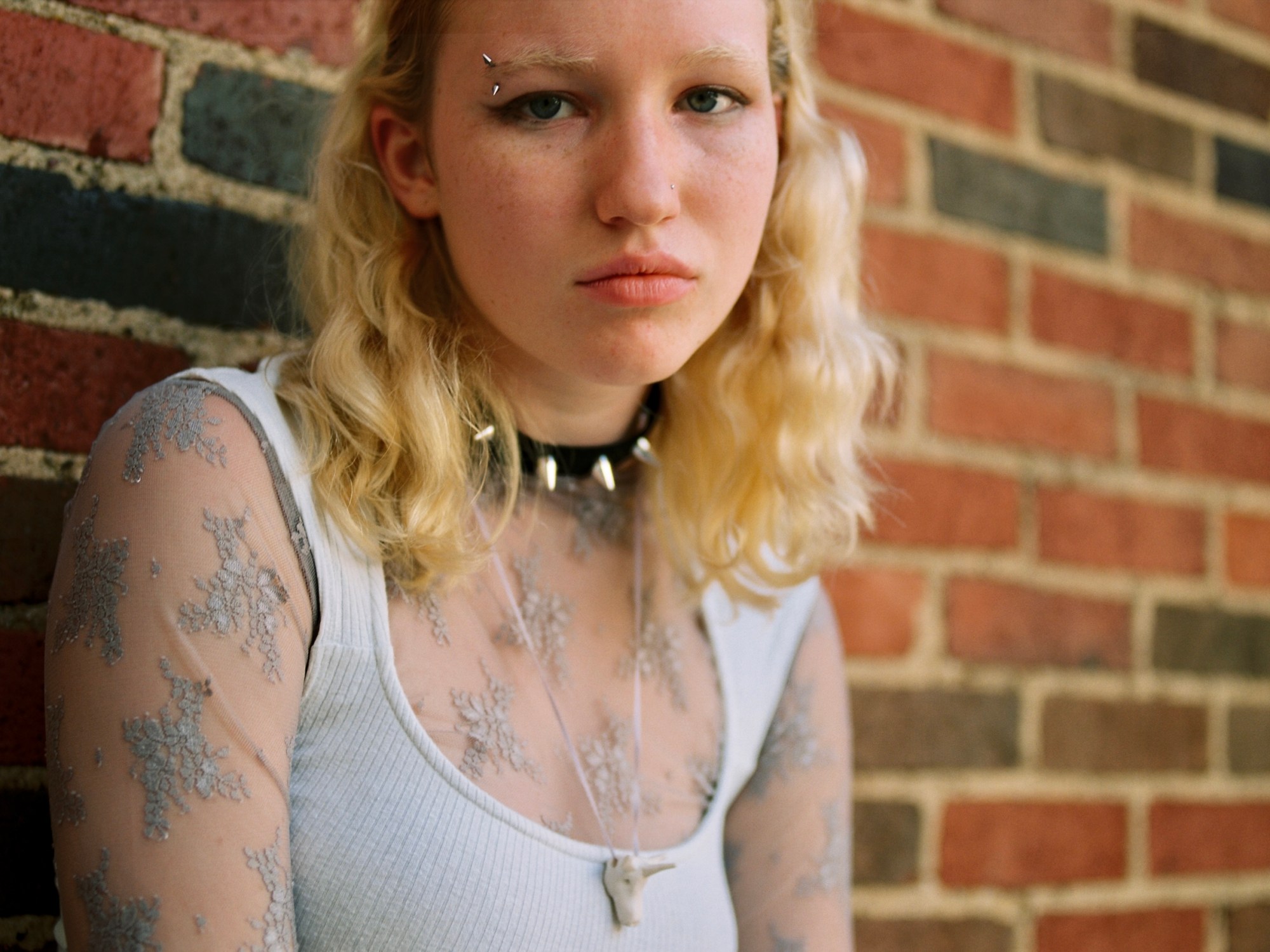 Photograph of a girl with a piercing in her bleached eyebrow, a studded choker, a unicorn charm necklace, a ribbed vest, and a sheer top standing against a brick wall