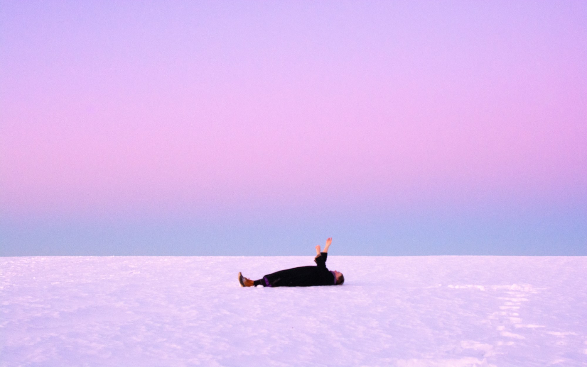 Photograph of a woman lying on the ice under a purple pink sky with her arms in the air