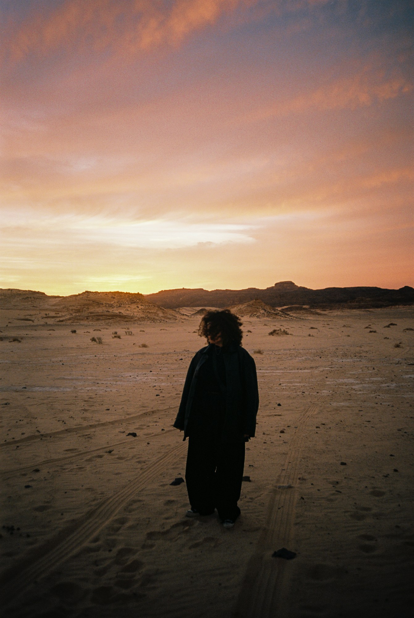 Photograph of someone standing in the middle of a desert amongst car tracks at sunset