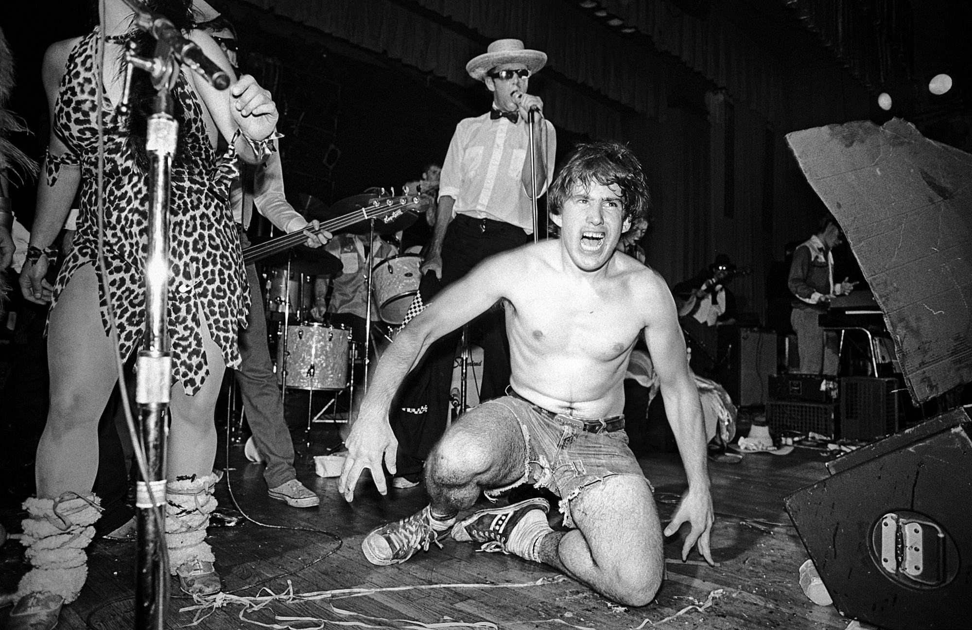 a topless billy goons in jean shorts on stage at a concert with a singer wearing cheetah print in the foreground and a dancer dressed like a cafe waiter in the background