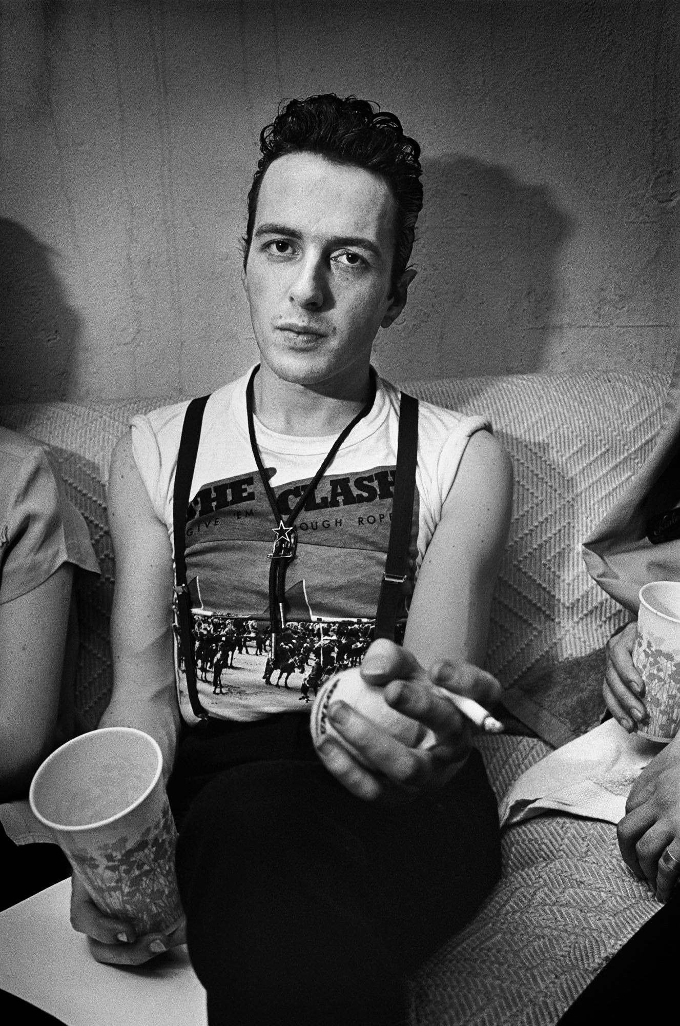 joe strummer smoking a cigarette wearing a clash t shirt looks into the camera. he's sat on a sofa.