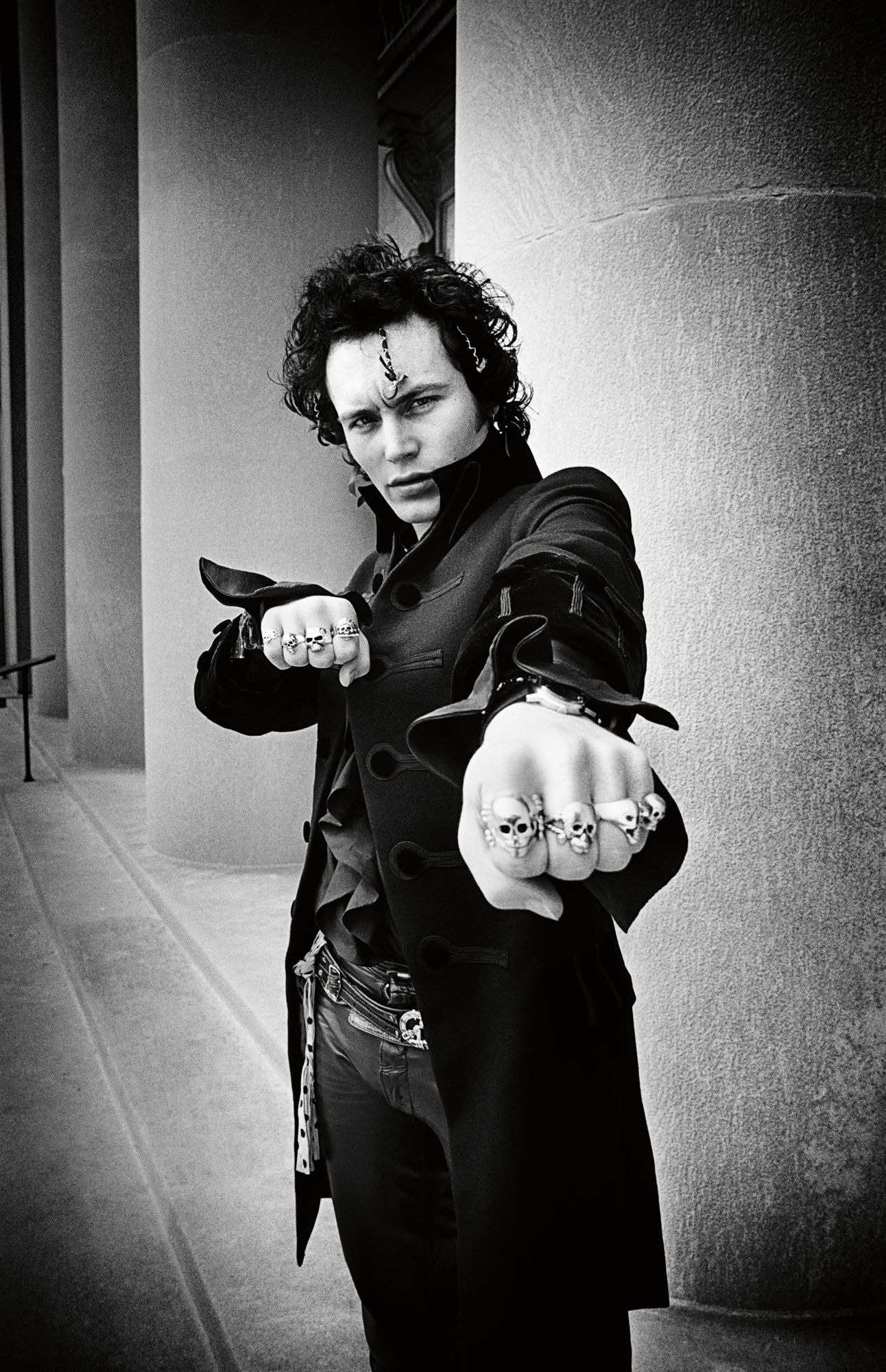 adam ant with knuckle rings and a leather jacket looks into the camera menacingly. he is posing in front of pillars.