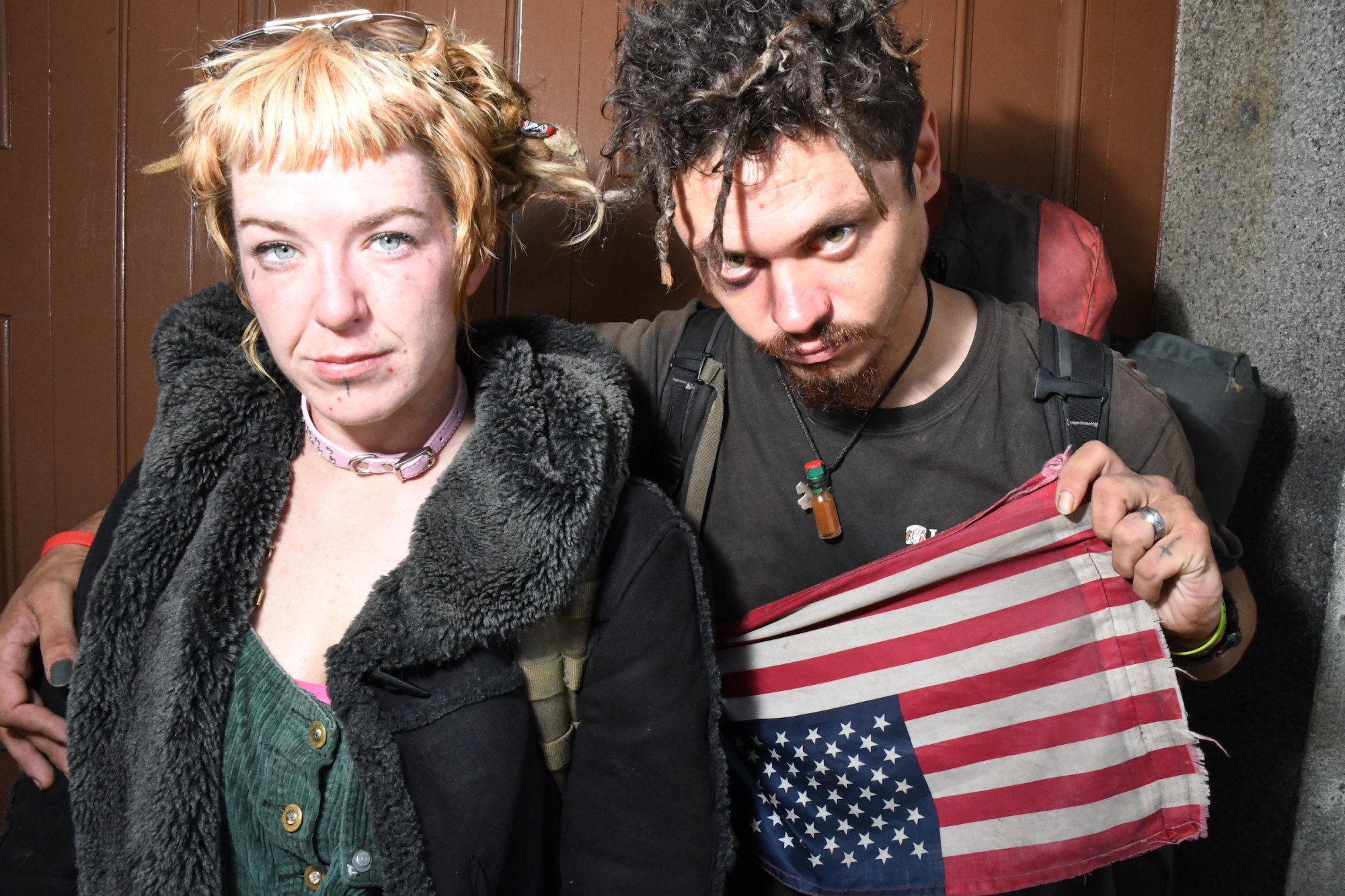 Photograph by Tom TBow Bowden of a couple with knotted hair who have been travelling on trains with backpacks. one holds an upside down american flag