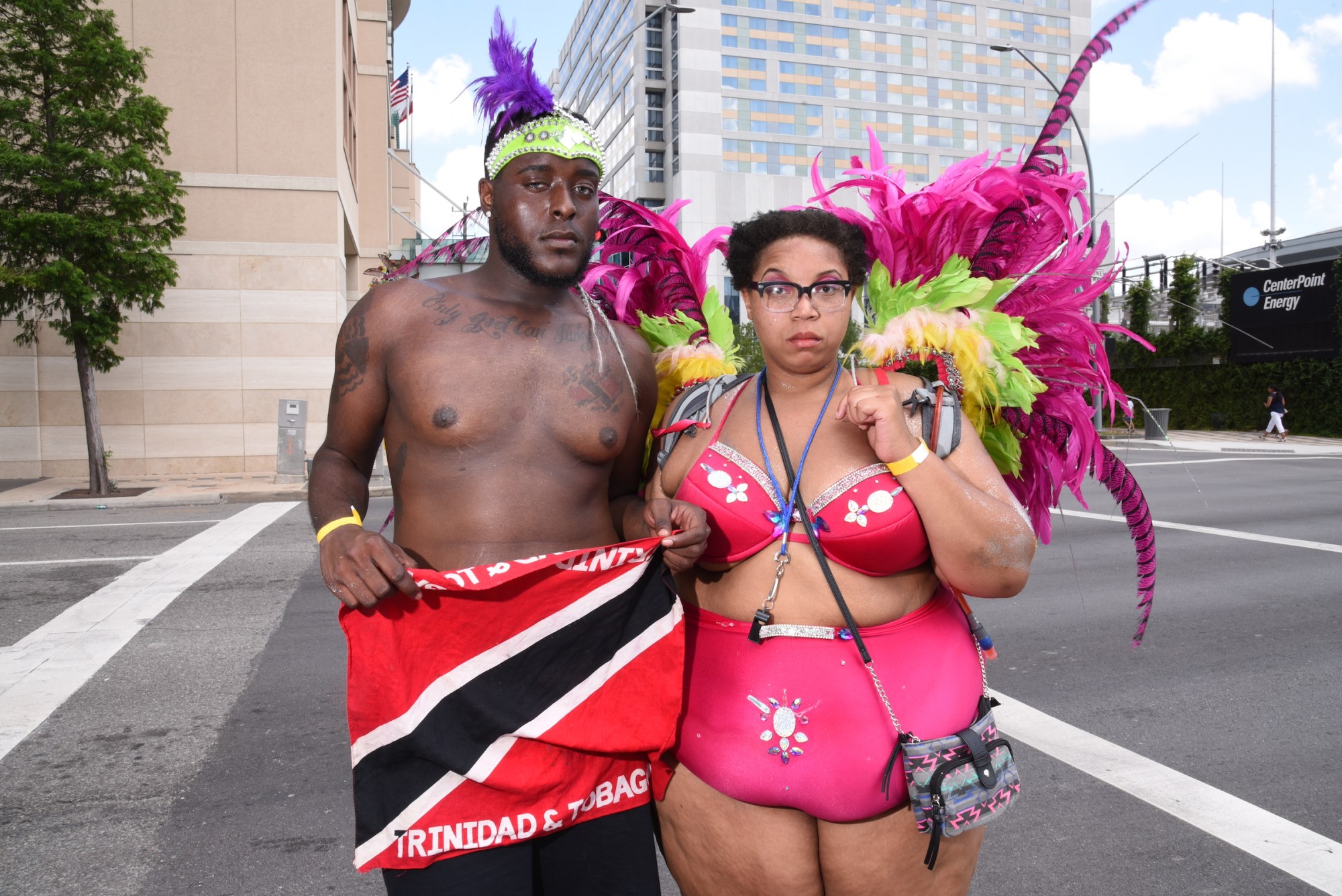 Photograph by Tom TBow Bowden of a couple, one shirtless the other wearing pink lingerie and feather wings at a festival. The former holds a Trinidad and Tobago flag.