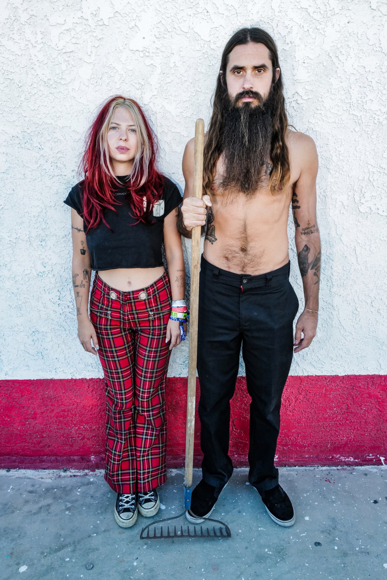 Photograph by Tom TBow Bowden of a couple with a woman with red and blonde hair and tartan trousers and a shirtless man with long hair and holding a rake.