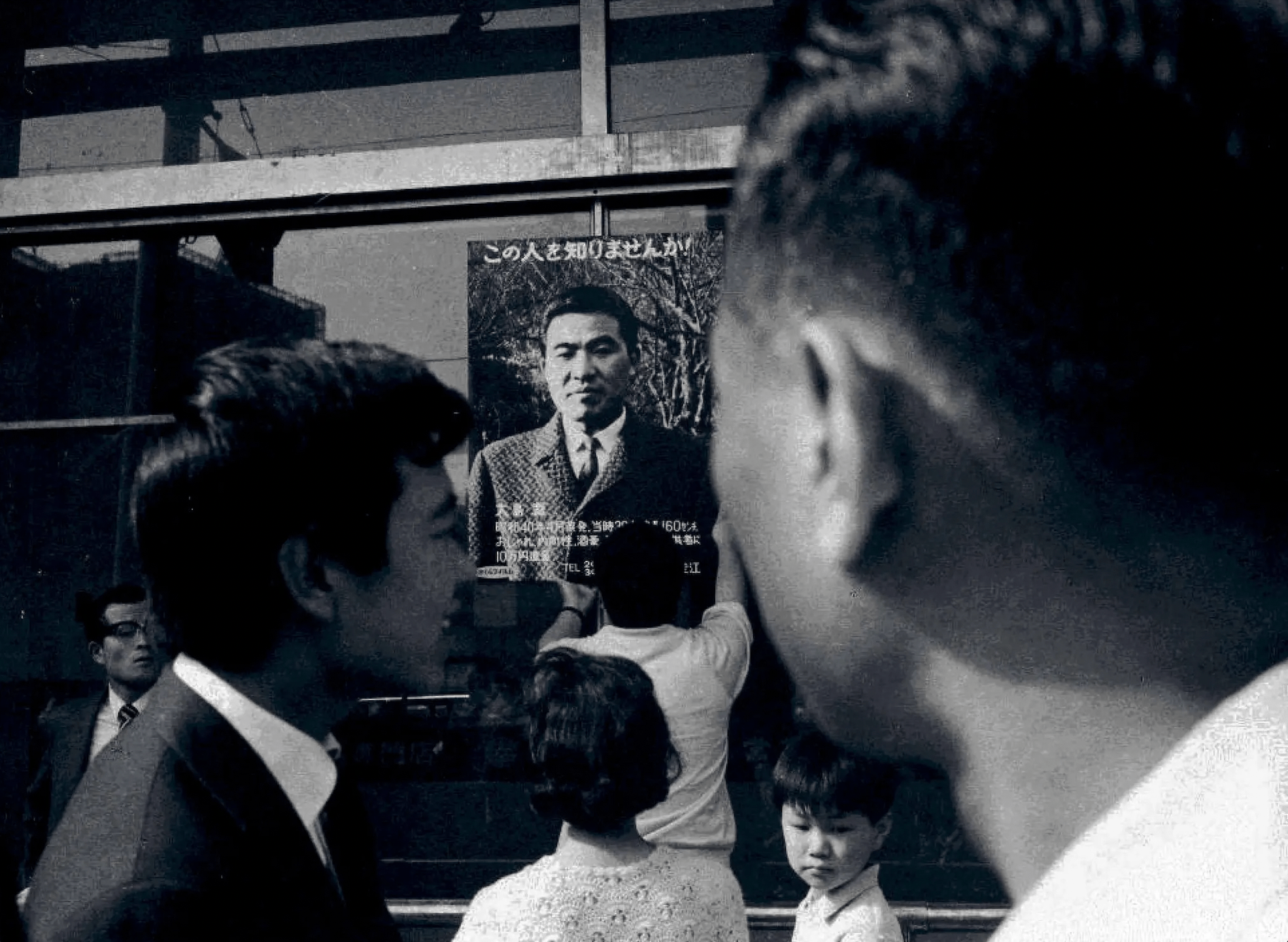 Black-and-white image of crowds walking in front of a missing poster of a man