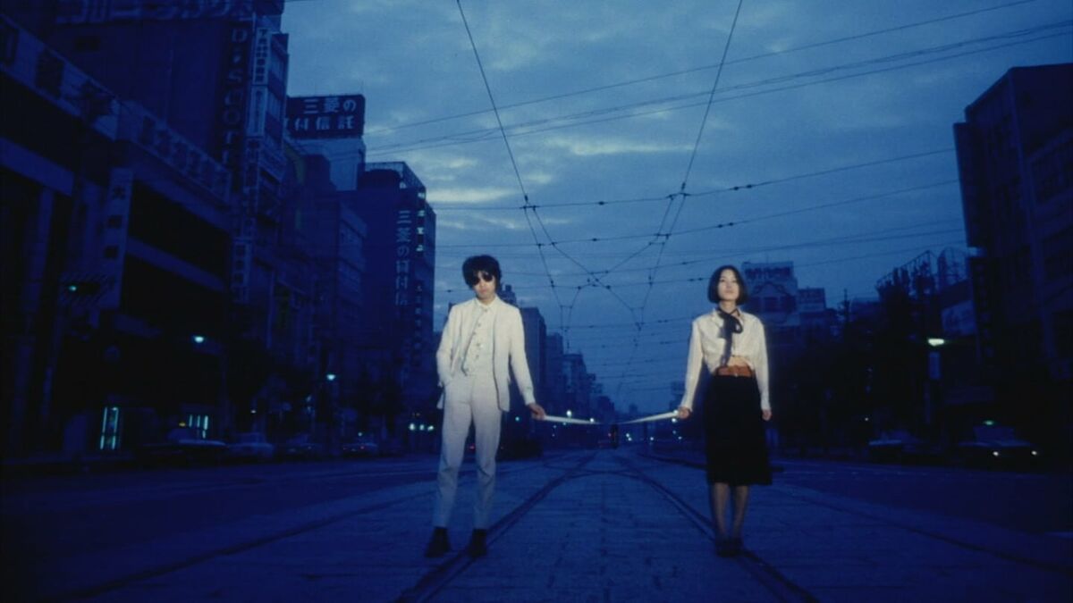 A couple stand parallel to each other on a blue-lit cityscape