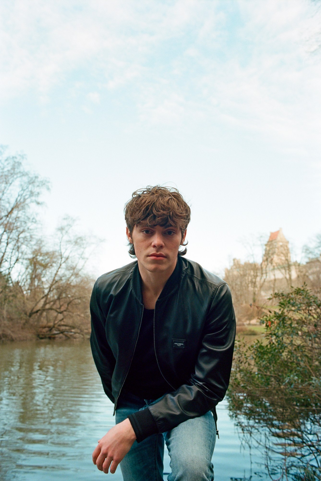 jack champion wearing a black denim jacket and blue jeans by the water in central park