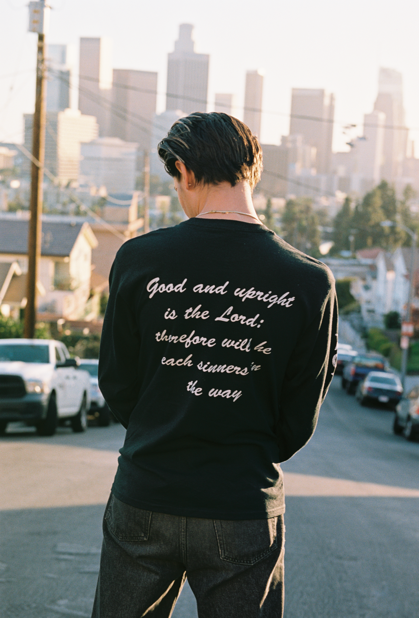 a model stands with their back to the camera on a long LA street; a bible verse appears to be on the back of his shirt