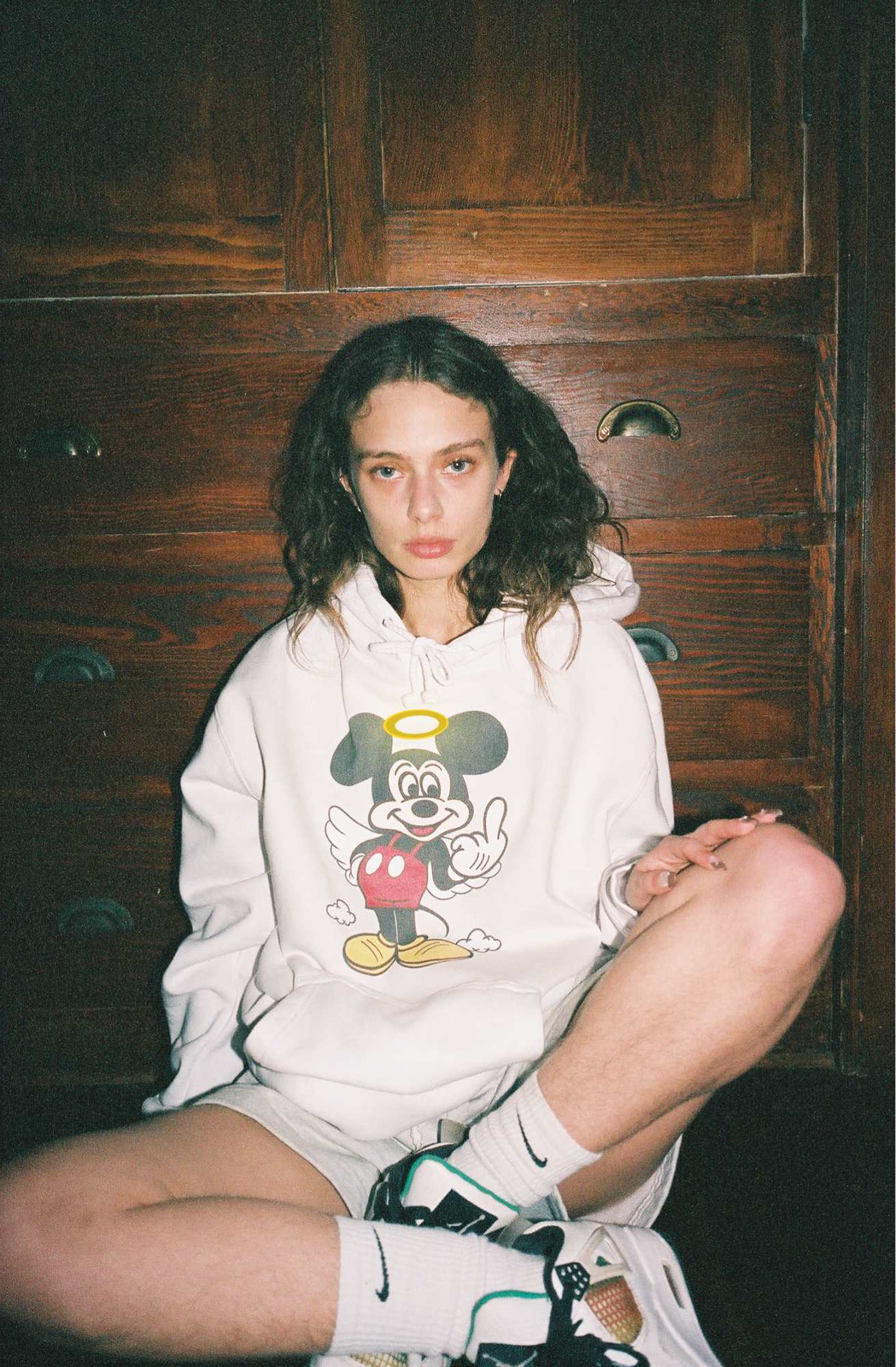 a girl sits cross-legged in white shorts and a white hoodie on which mickey mouse is swearing