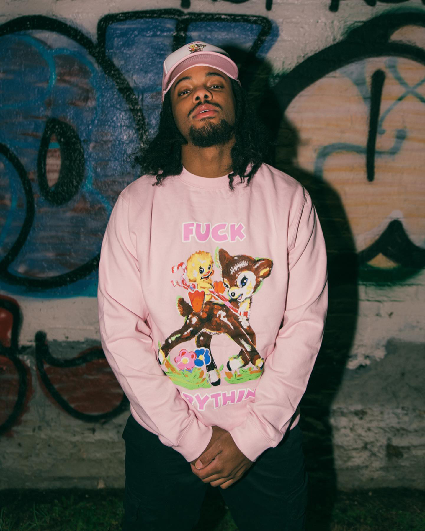 a man stands against a graffitied wall wearing a pink sweater with a cute bambi image and the words FUCK EVERYTHING