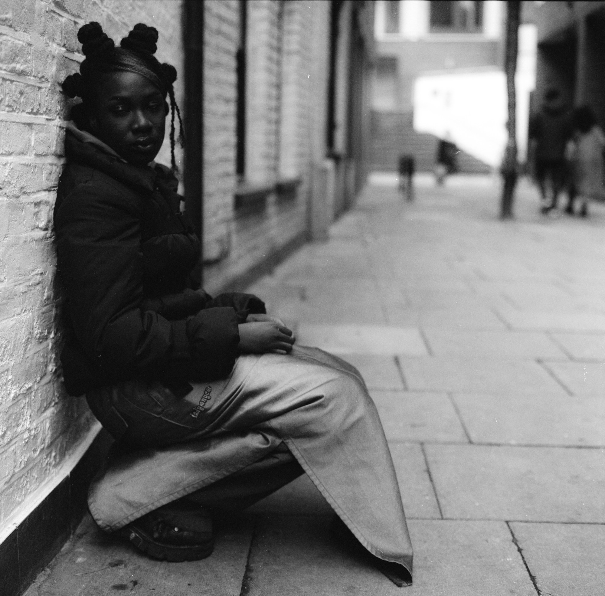 the actor vivian oparah wearing a puffer jacket with her hair in buns. she is crouching against a brick wall wearing a skirt with a wide seam split.