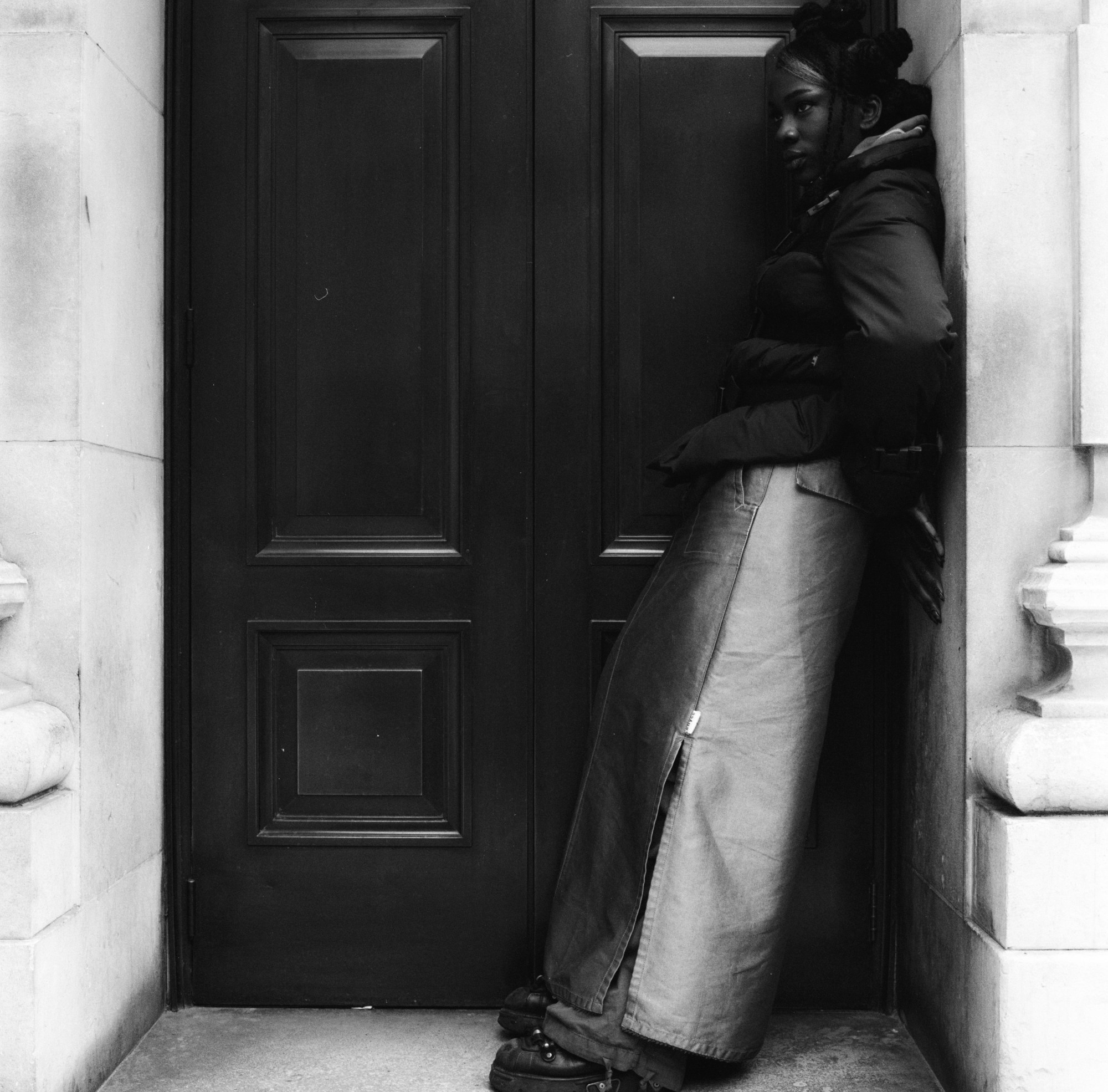 the actor vivian oparah wearing a puffer jacket with her hair in buns. she is standing in a door way, wearing a puffer jacket and a skirt with a high seam split, she is looking off camera.