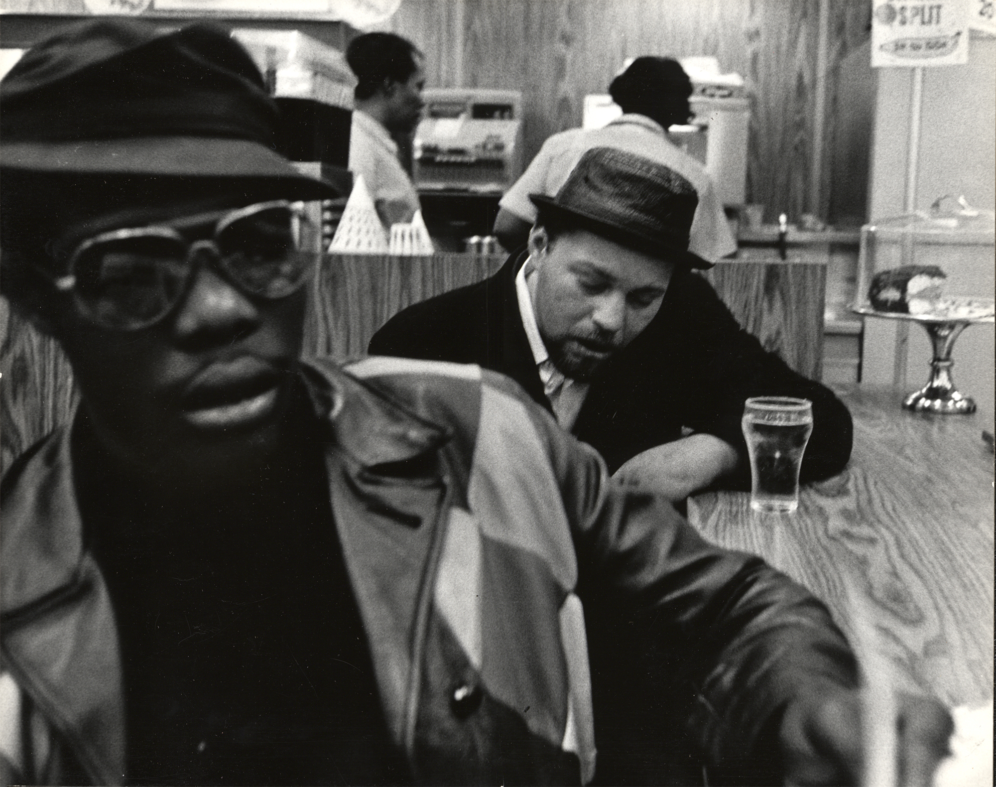 a stylish black man in a pattterned leather jacket sits at the bar of a cafe, in the background, a man in a trilby hat drinking a small pint.